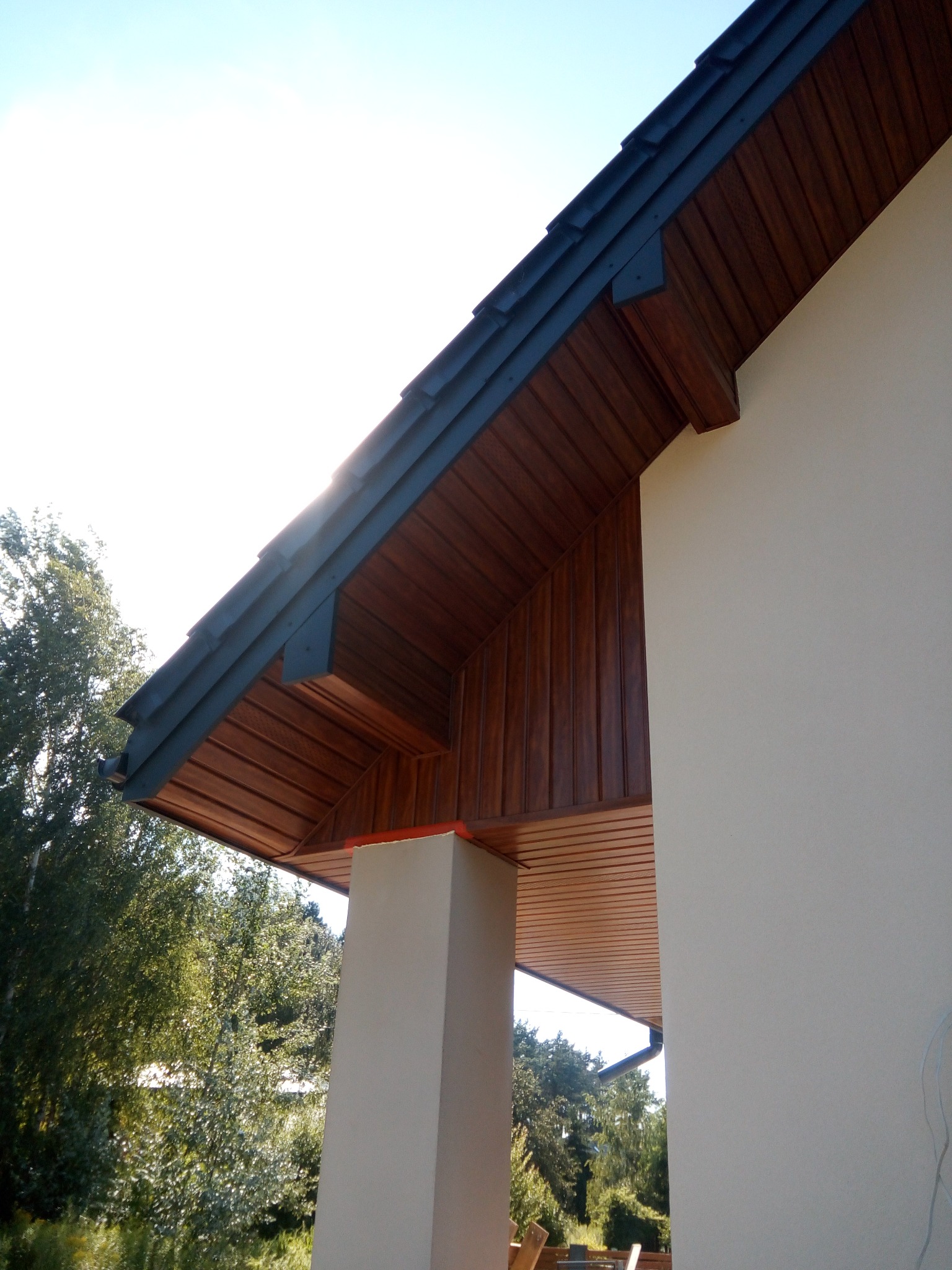 Drewniana podbitka dachowa z ciemnymi elementami wykończeniowymi, widok z dołu na fragment elewacji budynku i zieleń w tle.