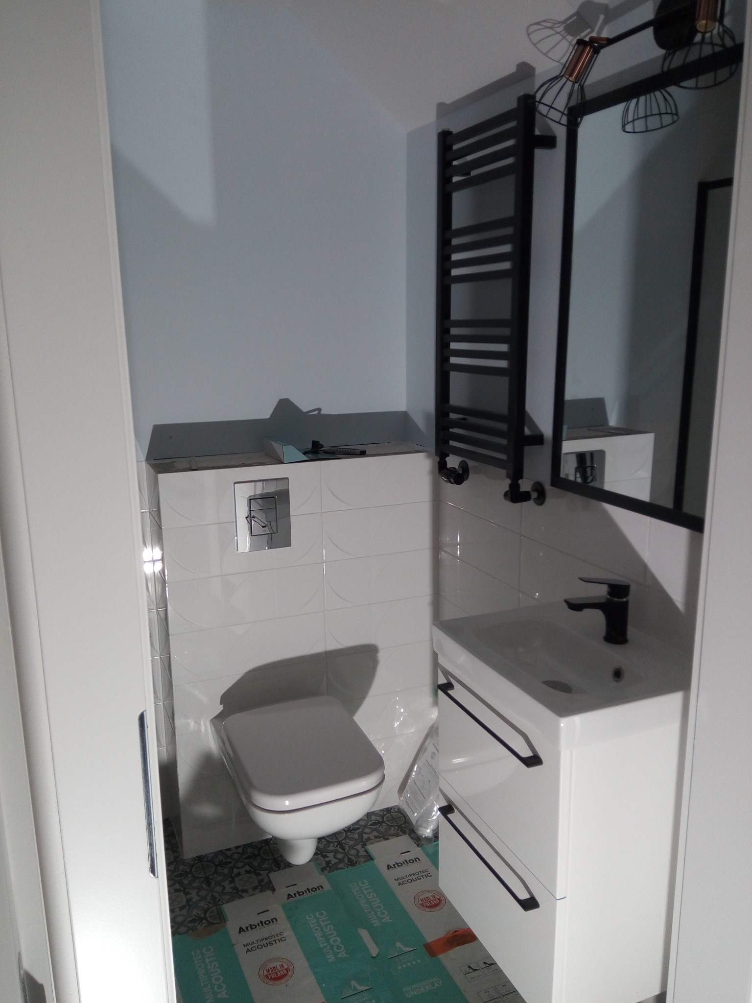 Nowoczesna łazienka po remoncie: białe płytki, wisząca toaleta, umywalka z szafką, czarny grzejnik drabinkowy i lustro z oświetleniem, widoczne kartony podkładu akustycznego 'Arbiton'.