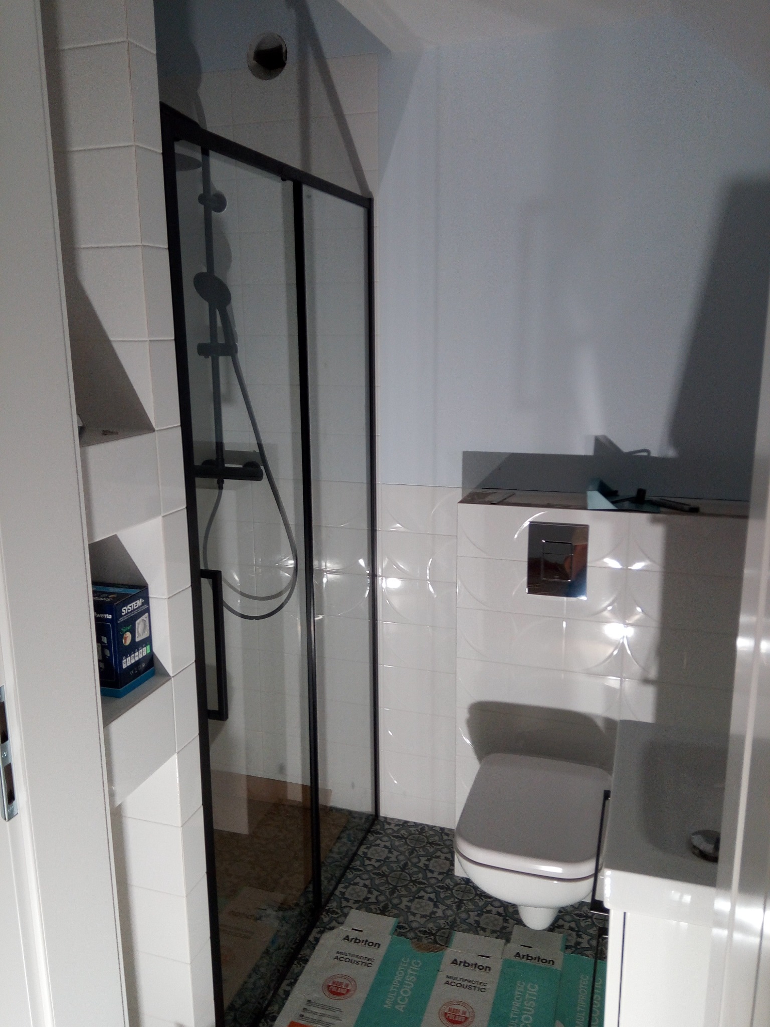 Nowoczesna łazienka po remoncie: białe płytki z delikatnym wzorem, kabina prysznicowa z czarną ramą, toaleta i umywalka, widoczne podkłady pod panele akustyczne na podłodze.