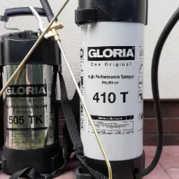 Dwa opryskiwacze ciśnieniowe Gloria, modele 505 TK i 410 T, stojące na czerwonej kostce brukowej przy ścianie budynku.