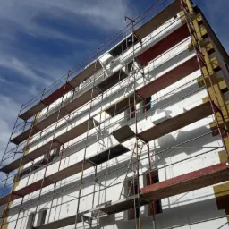 Wielopiętrowy budynek w trakcie ocieplania, widok z dołu na rusztowania i białe płyty izolacyjne z widocznymi oknami, fragmenty żółtej wełny mineralnej na krawędziach, błękitne niebo z chmurami w tle.