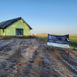 www.domyzkeramzytu.pl-Krotoszyn - Nowoczesny, parterowy dom z zieloną elewacją i ciemną dachówką, częściowo wykończony, z widocznym banerem reklamowym firmy budowlanej specjalizującej się w budowie domów energooszczędnych,...