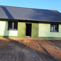 www.domyzkeramzytu.pl-Krotoszyn - Niedokończony, jednopiętrowy dom z zieloną elewacją i ciemnoszarą dachówką, widoczne ślady opon na błotnistym terenie przed budynkiem.