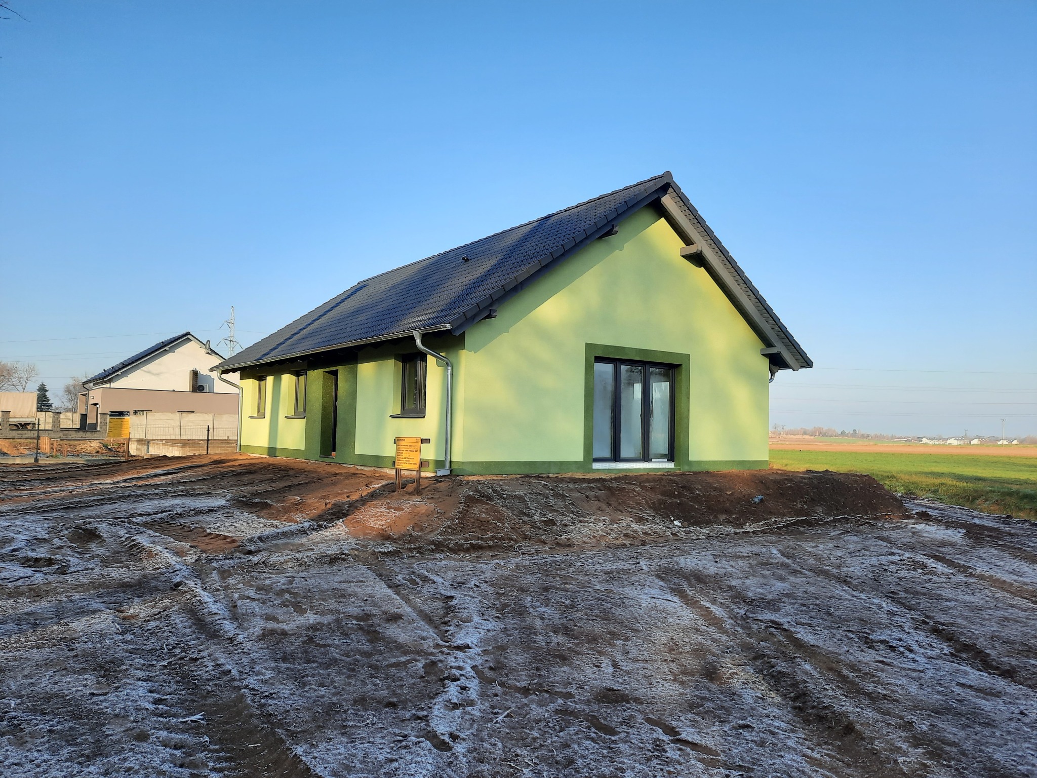 Nowo wybudowany, parterowy dom z zieloną elewacją i ciemną dachówką, otoczony ziemią z delikatnym oszronieniem, w tle widoczne pole i błękitne niebo.