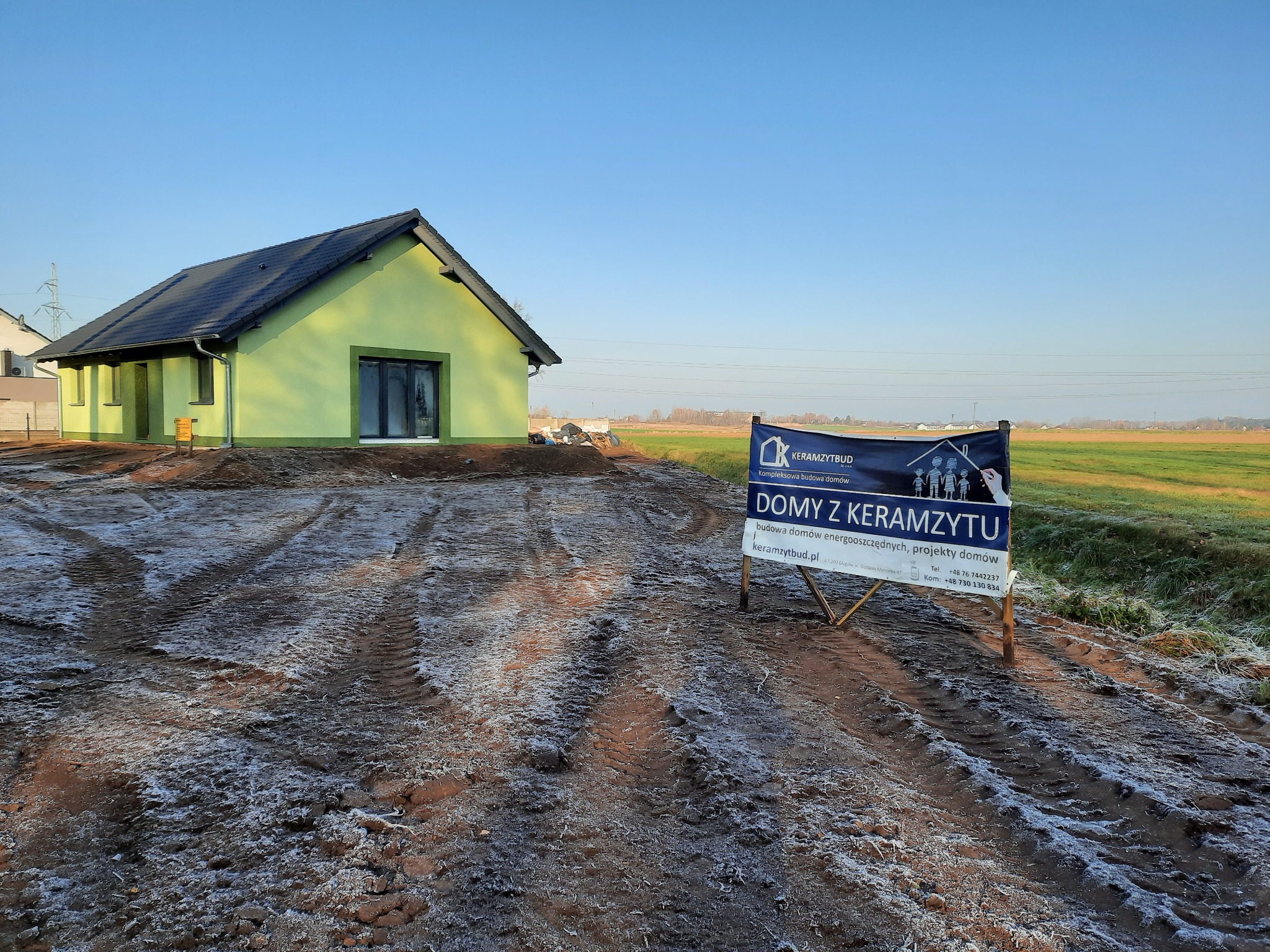 Nowoczesny, parterowy dom z zieloną elewacją i ciemną dachówką, częściowo wykończony, z widocznym banerem reklamowym firmy budowlanej specjalizującej się w budowie domów energooszczędnych,...