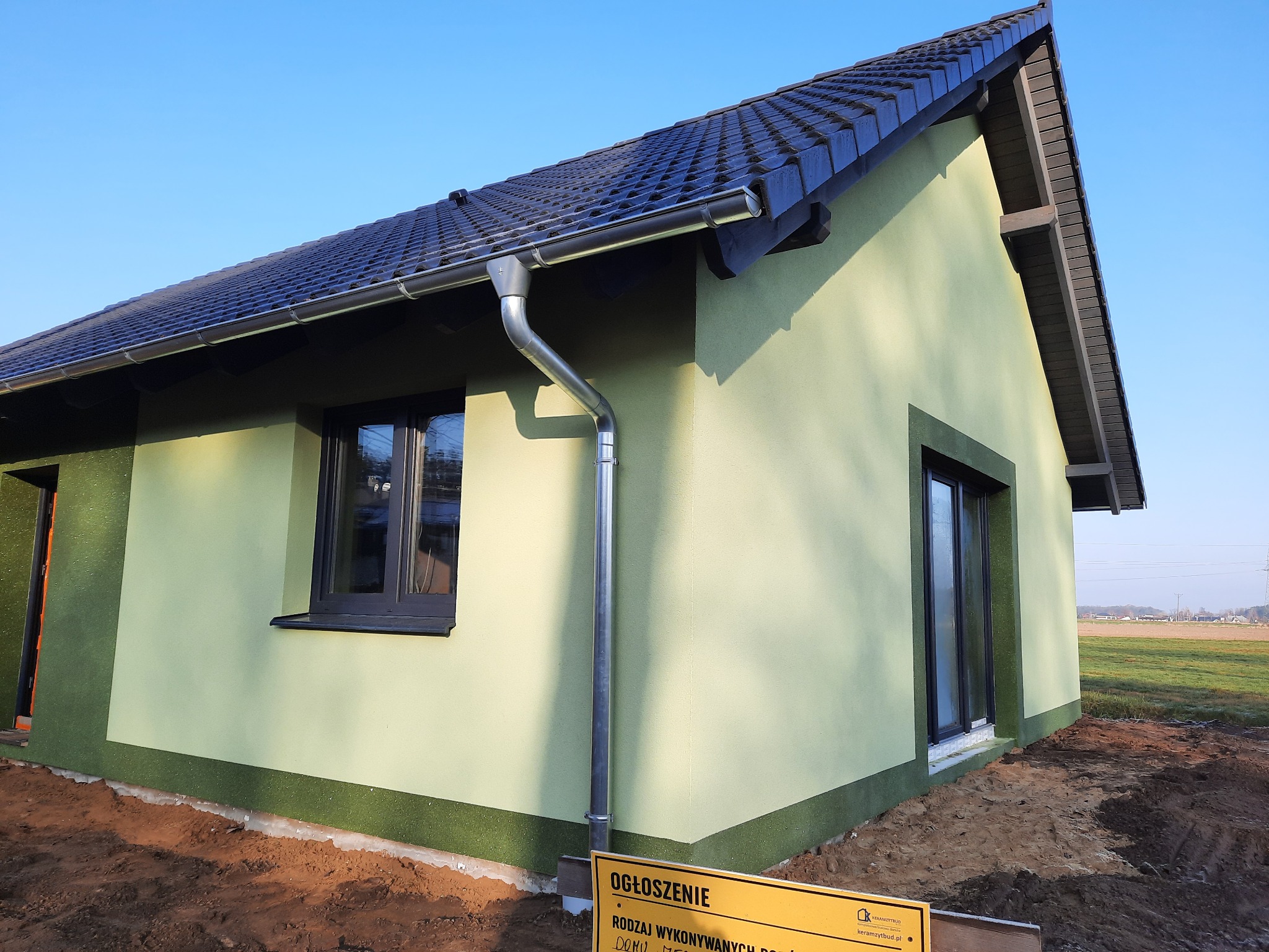 Nowo wybudowany, parterowy dom z jasnozieloną elewacją, ciemną dachówką i metalową rynną, na działce z częściowo wyrównanym terenem.