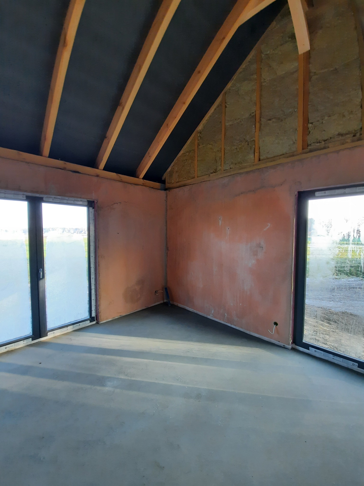 Wnętrze domu w stanie surowym zamkniętym: widoczne ocieplenie wełną mineralną poddasza, ściany pokryte tynkiem cementowo-wapiennym, betonowa posadzka oraz duże okna przesuwne.