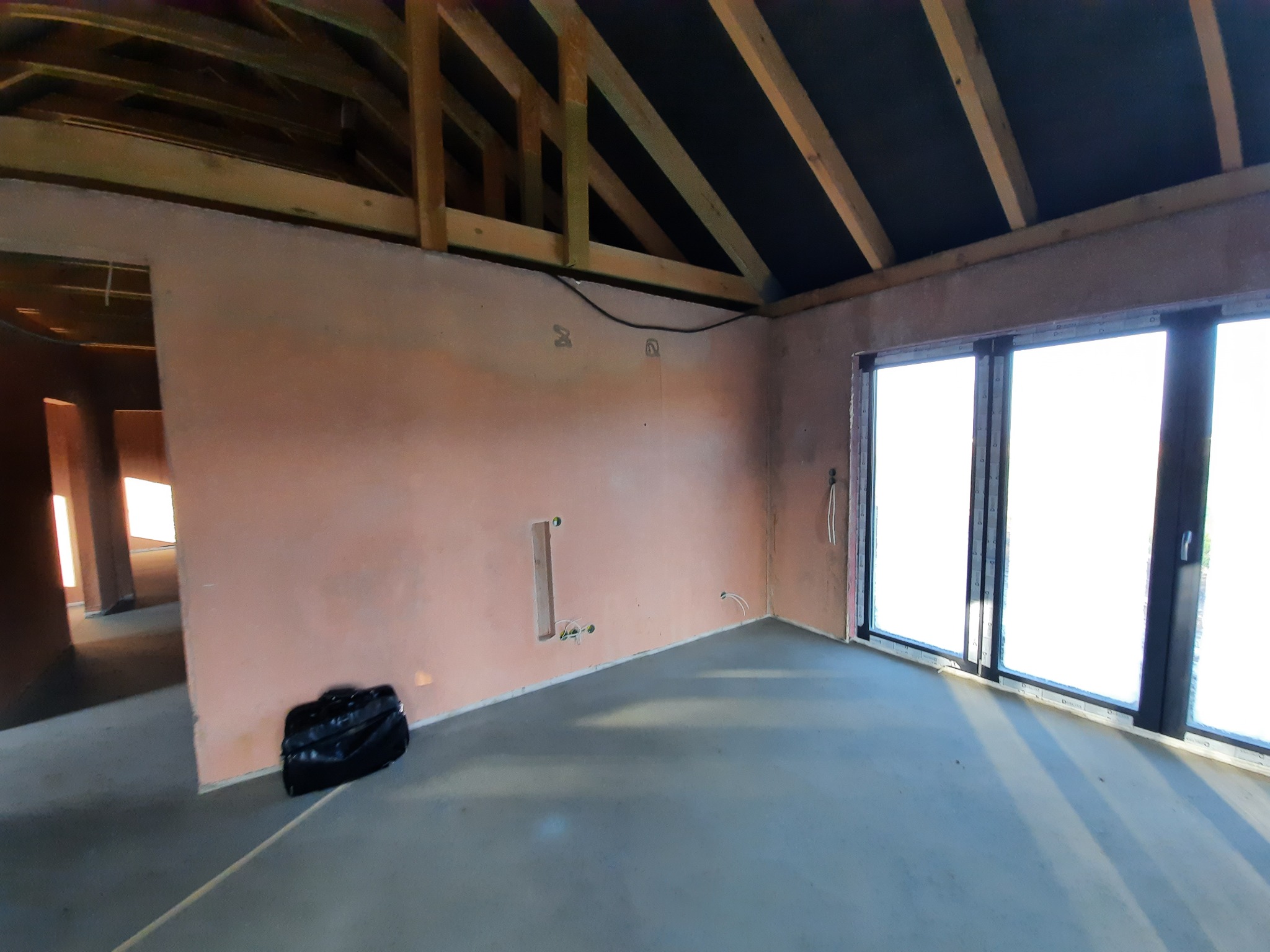 Wnętrze surowego domu w trakcie budowy: ściany otynkowane na różowo, widoczne instalacje elektryczne, drewniana konstrukcja dachu i duże okno balkonowe z czarnymi ramami. Na podłodze widać wylany...