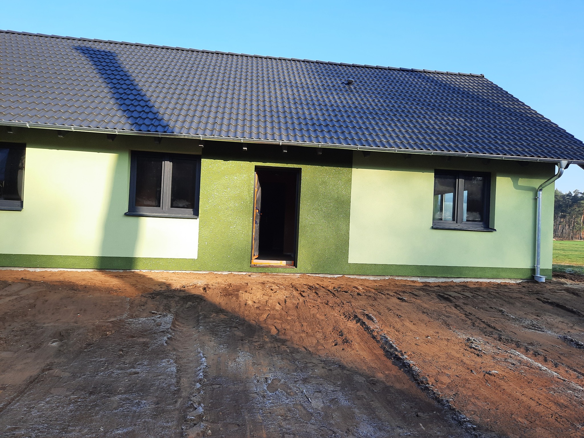 Niedokończony, jednopiętrowy dom z zieloną elewacją i ciemnoszarą dachówką, widoczne ślady opon na błotnistym terenie przed budynkiem.