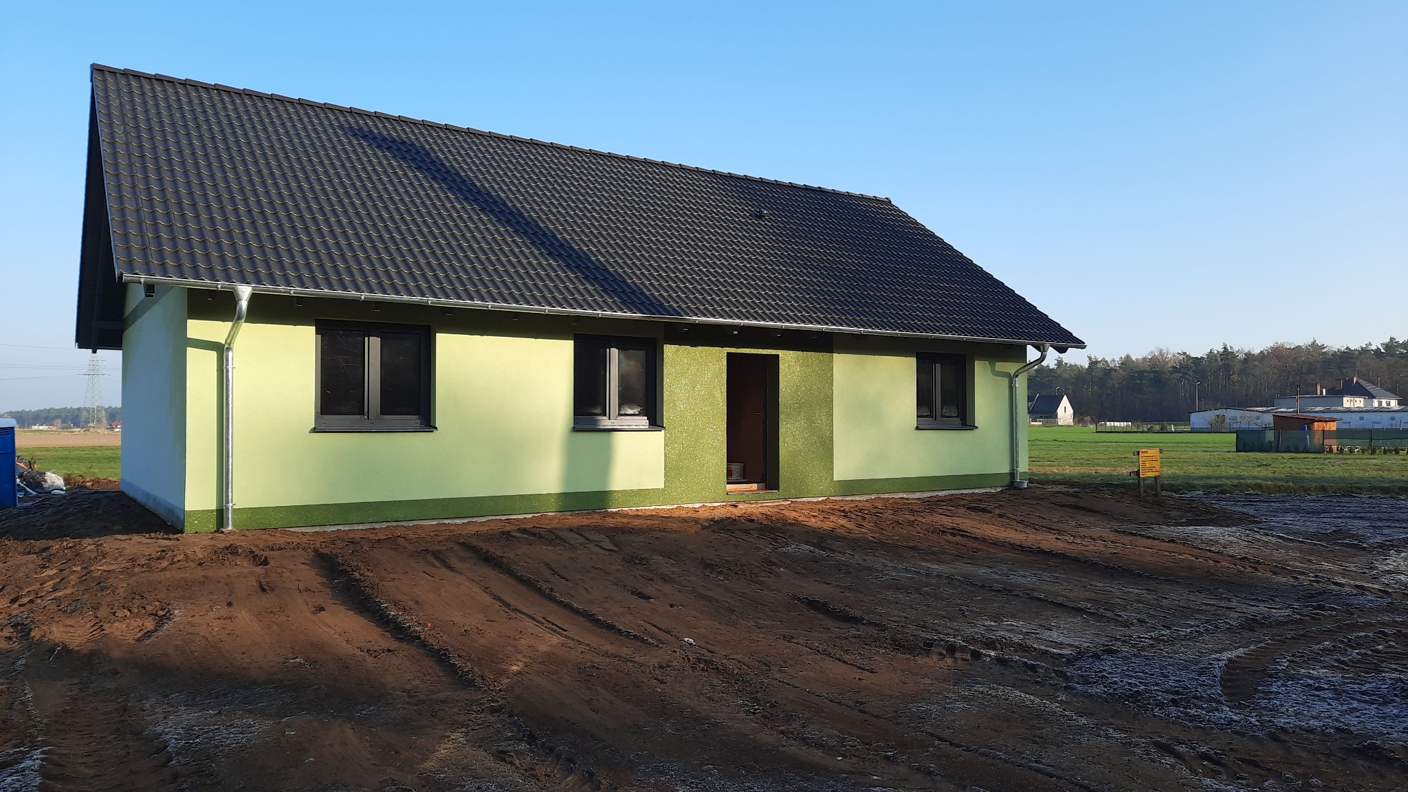 Niedokończony parterowy budynek z zielonymi ścianami i ciemną dachówką, widoczne ślady opon na ziemi, w tle łąka z zabudowaniami.
