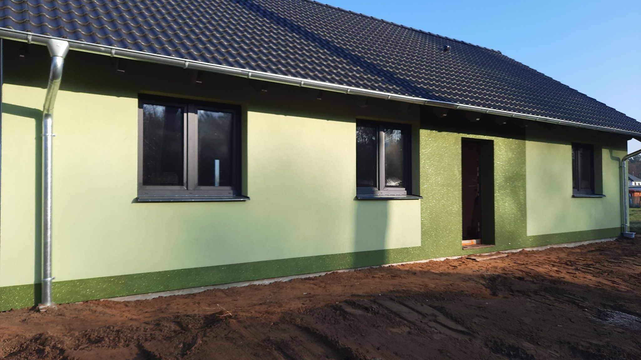 Świeżo otynkowany dom jednorodzinny z czarną dachówką i grafitową stolarką okienną, widoczne orynnowanie i wstępnie wyrównany teren wokół budynku.