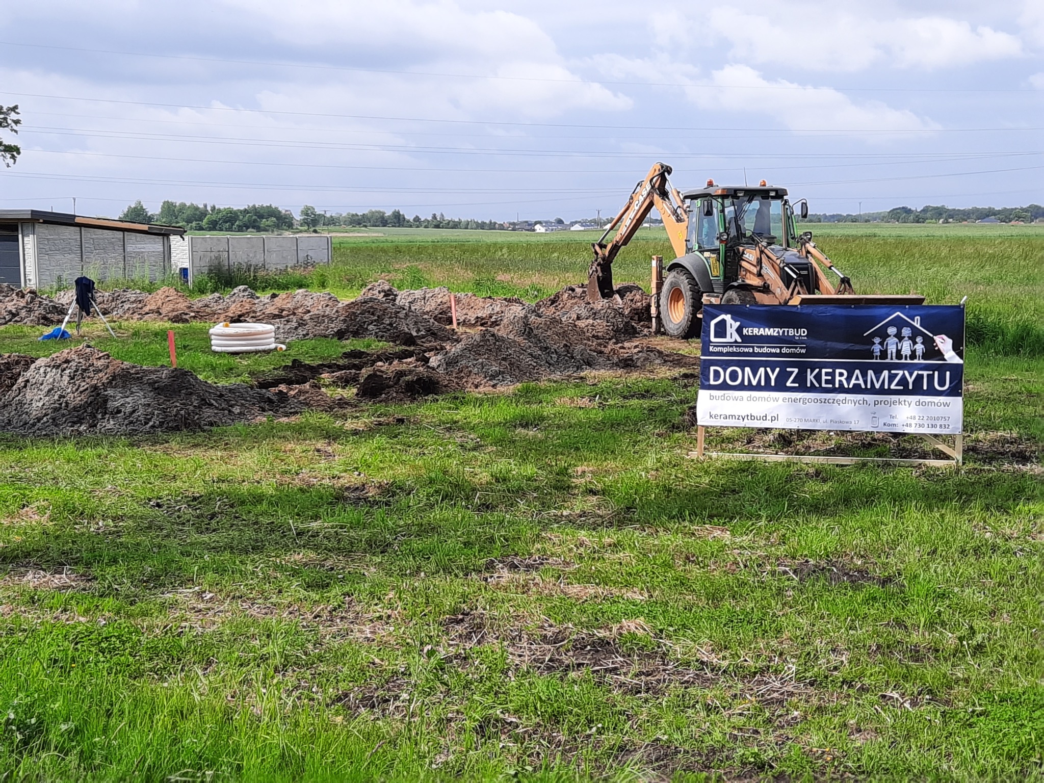Żółto-czarna koparko-ładowarka przygotowuje teren pod budowę domu, w tle widoczny znak reklamowy z napisem 'Domy z keramzytu', na pierwszym planie wykopana ziemia i białe rury.