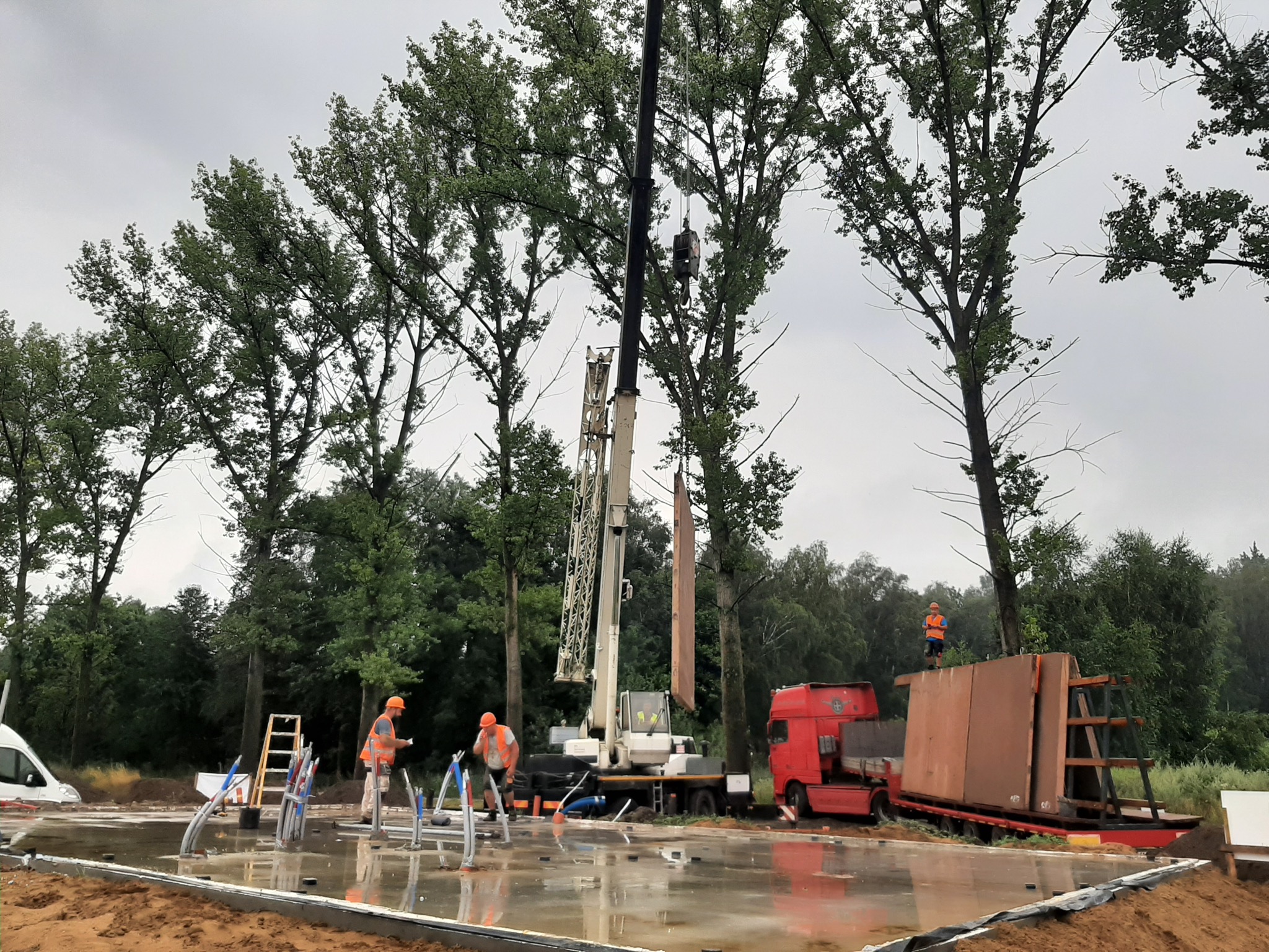 Montaż prefabrykowanych ścian domu na fundamencie z użyciem dźwigu, dwóch pracowników w kamizelkach odblaskowych i kaskach, oraz transportu na czerwonej ciężarówce. W tle drzewa i pochmurne niebo.