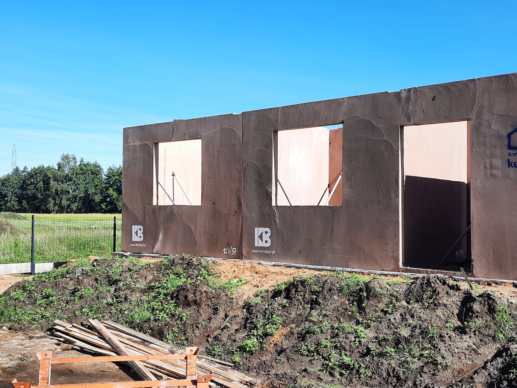 Surowy stan ścian zewnętrznych budynku z keramzytu z otworami okiennymi, widoczne logo firmy budowlanej na elewacji, ziemia i deski na pierwszym planie.