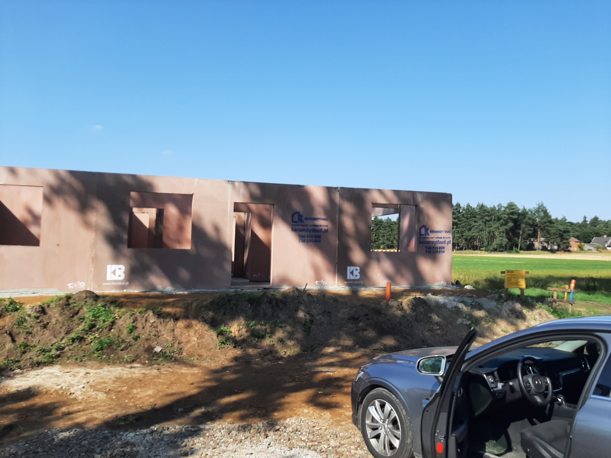 Budowa domu z keramzytu w stanie surowym otwartym, widoczne ściany z otworami okiennymi i drzwiowymi, logo firmy budowlanej na ścianie, samochód osobowy z otwartymi drzwiami na pierwszym planie...