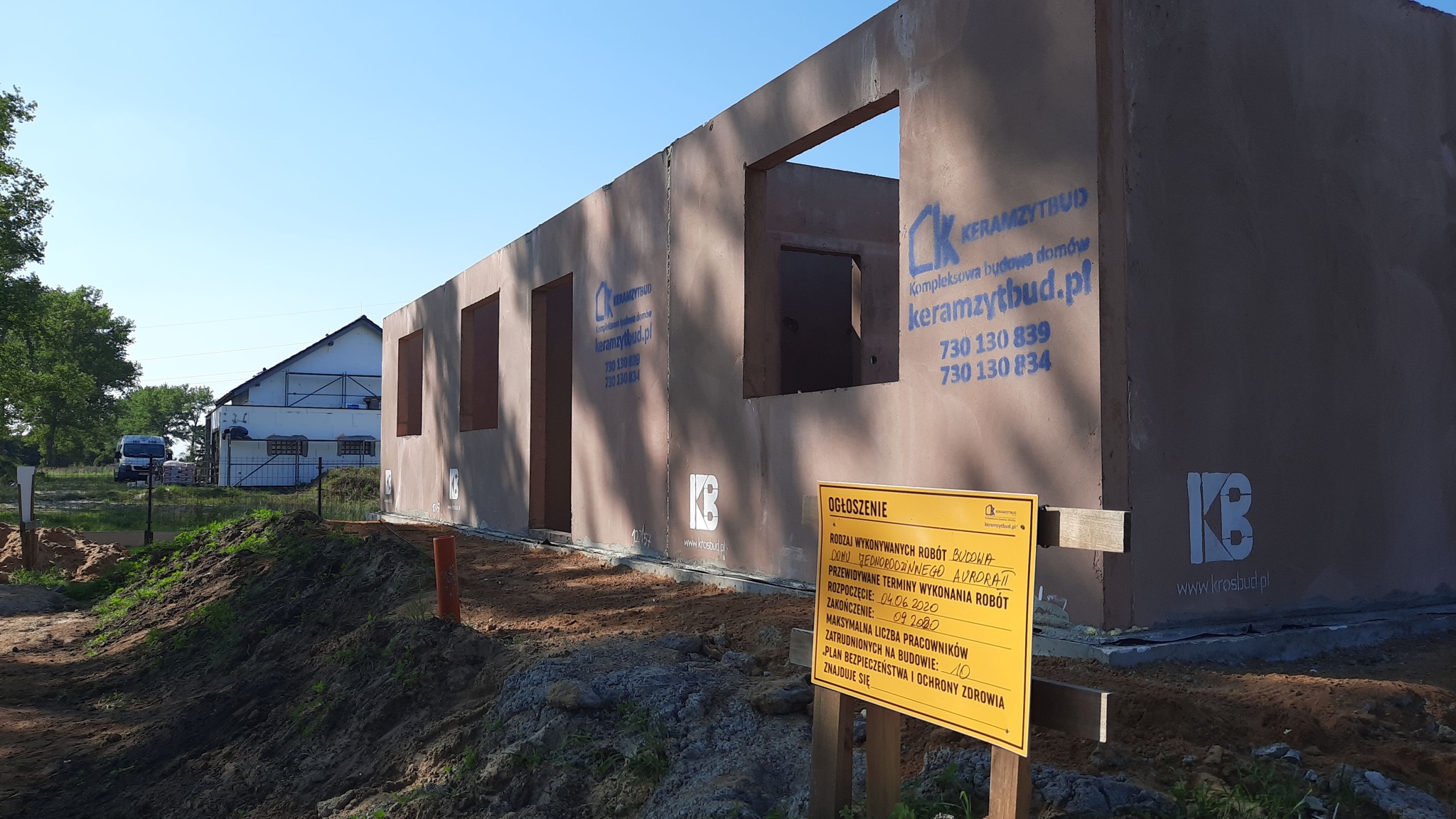 Surowy stan domu z keramzytu z widocznym ogłoszeniem budowlanym, informującym o rodzaju wykonywanych robót: budowa domu jednorodzinnego Aurora. W tle zabudowania gospodarcze i częściowo zalesiony...