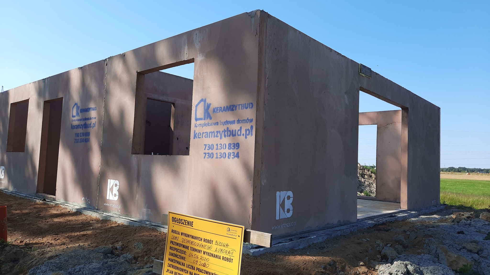 Surowy stan domu z keramzytu z widocznymi otworami okiennymi i drzwiowymi, na ścianach naniesione logo firmy budowlanej, widoczna tablica informacyjna budowy.