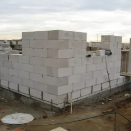 Wznoszenie ścian parteru domu z białych bloczków betonowych na fundamencie, widoczne elementy szalunku i rury wentylacyjne w murze, pochmurne niebo w tle.