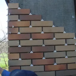 Fragment elewacji budynku w trakcie montażu cegieł klinkierowych, widoczne plastikowe krzyżyki dystansowe, siatka podtynkowa i częściowo zasłonięte okno z brązowymi roletami.