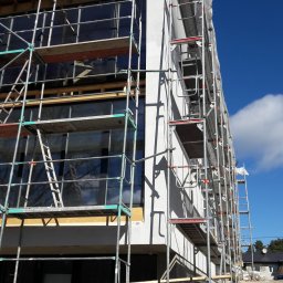 OLKO-NOME - Elewacja domu w trakcie prac wykończeniowych, widoczne rusztowania i fragment ocieplenia. Błękitne niebo w tle.