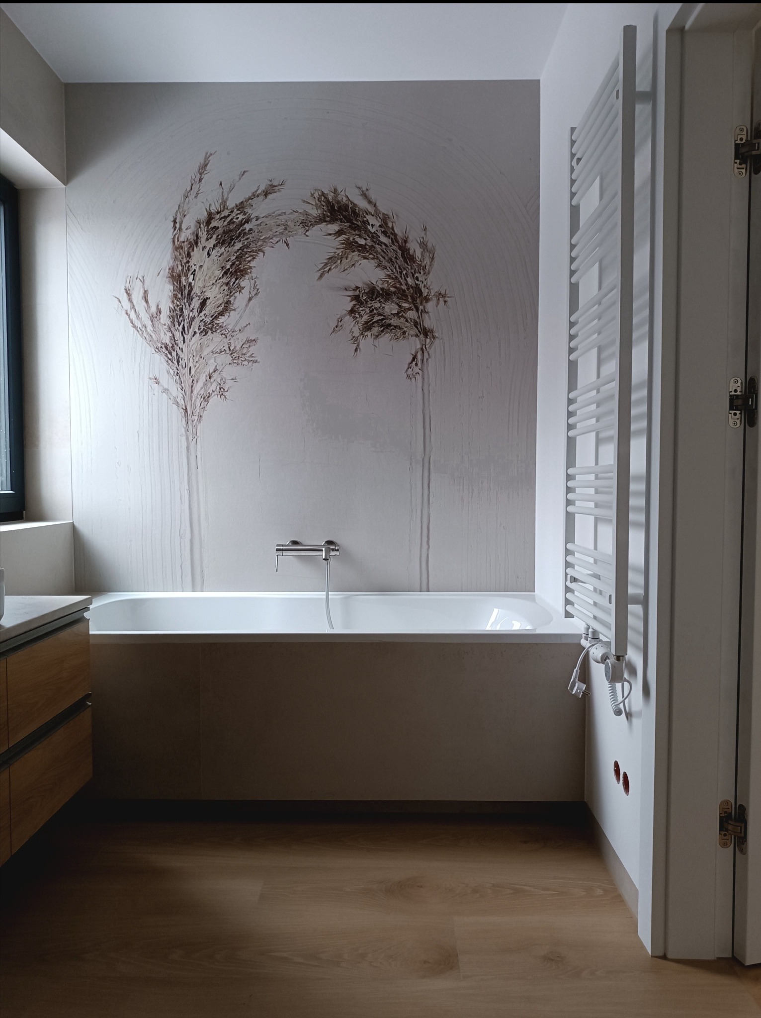 Nowoczesna łazienka z wanną obudowaną płytami, ozdobną tapetą z motywem roślinnym, drewnopodobną podłogą i grzejnikiem drabinkowym w kolorze białym.