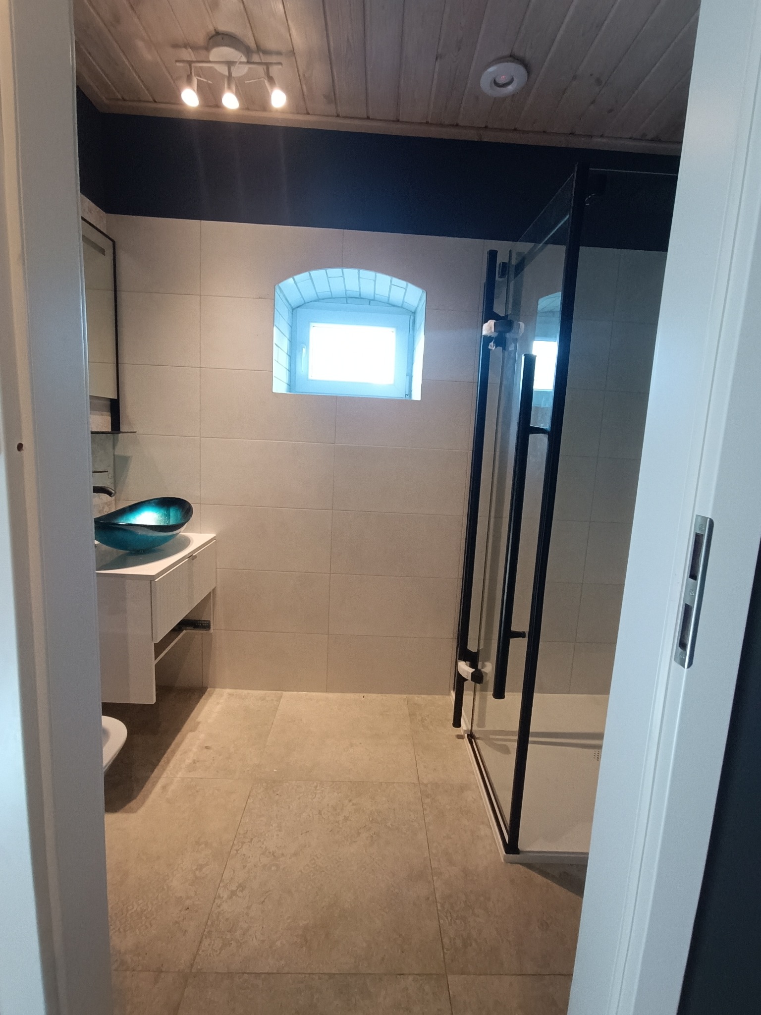 Nowoczesna łazienka z kabiną prysznicową typu walk-in, umywalką w kształcie misy i drewnianym sufitem. Okno z łukiem nadaje wnętrzu unikalny charakter.