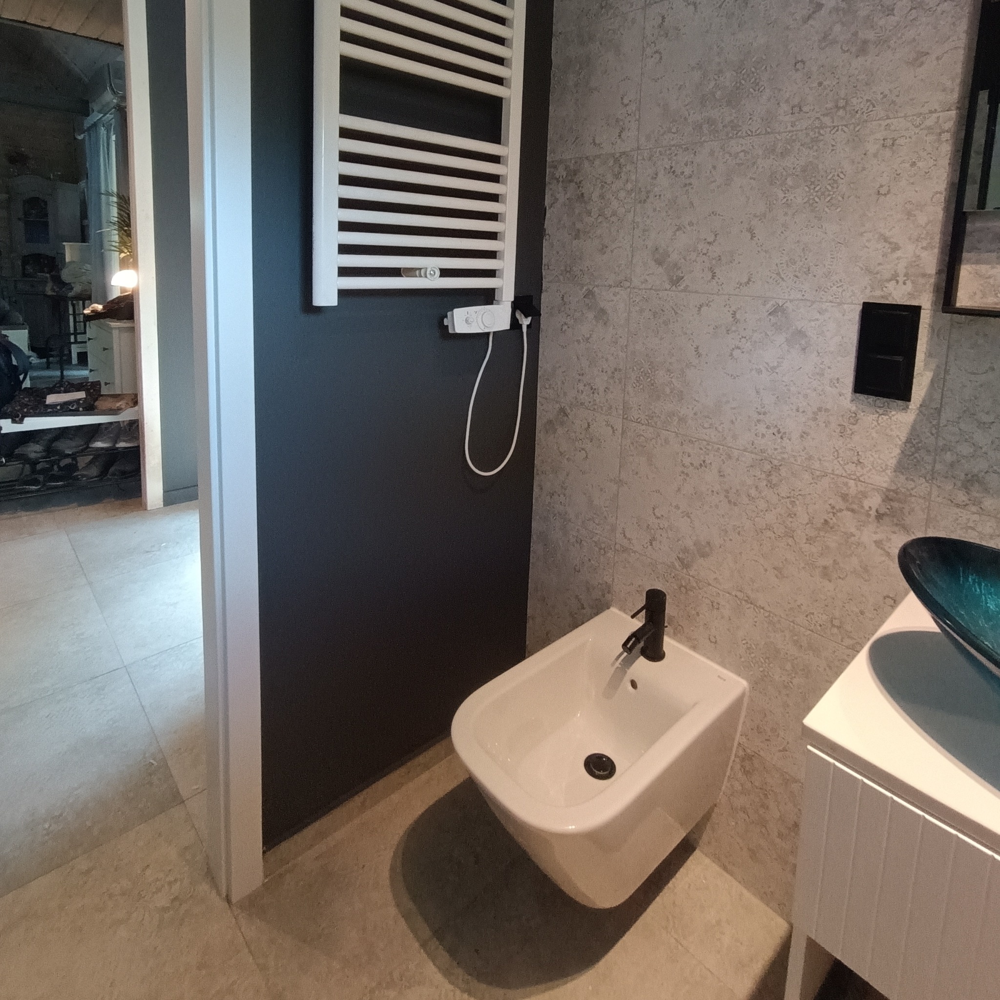 Nowoczesna łazienka z bidetem, grzejnikiem drabinkowym i umywalką z szafką. Szare płytki na ścianie i podłodze. Widok na inne pomieszczenie przez otwarte drzwi.