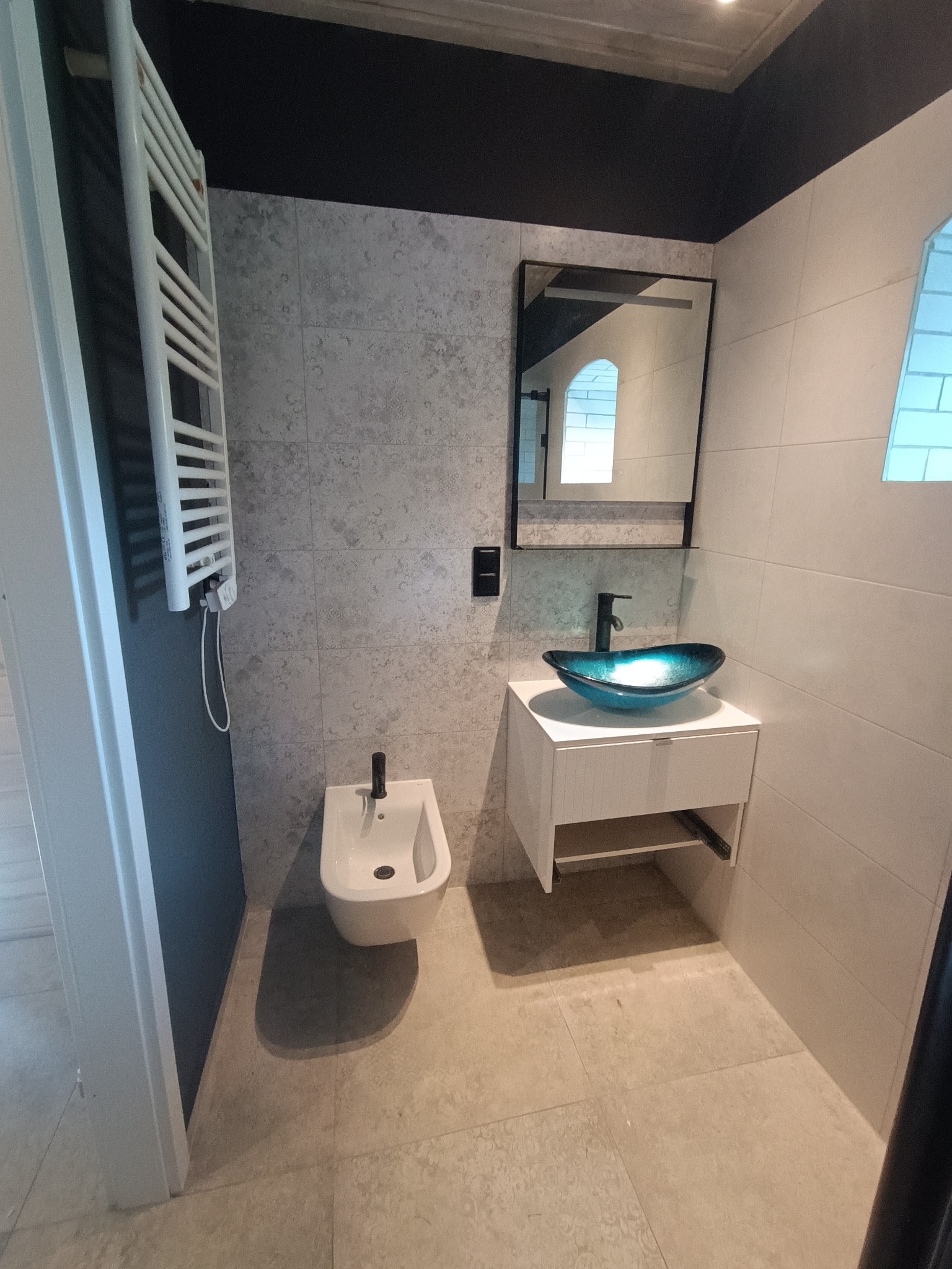 Nowoczesna łazienka z bidetem, grzejnikiem drabinkowym, umywalką z szafką i lustrem. Płytki w odcieniach szarości i beżu. Ciemny sufit. Wykończenie z dbałością o detale.