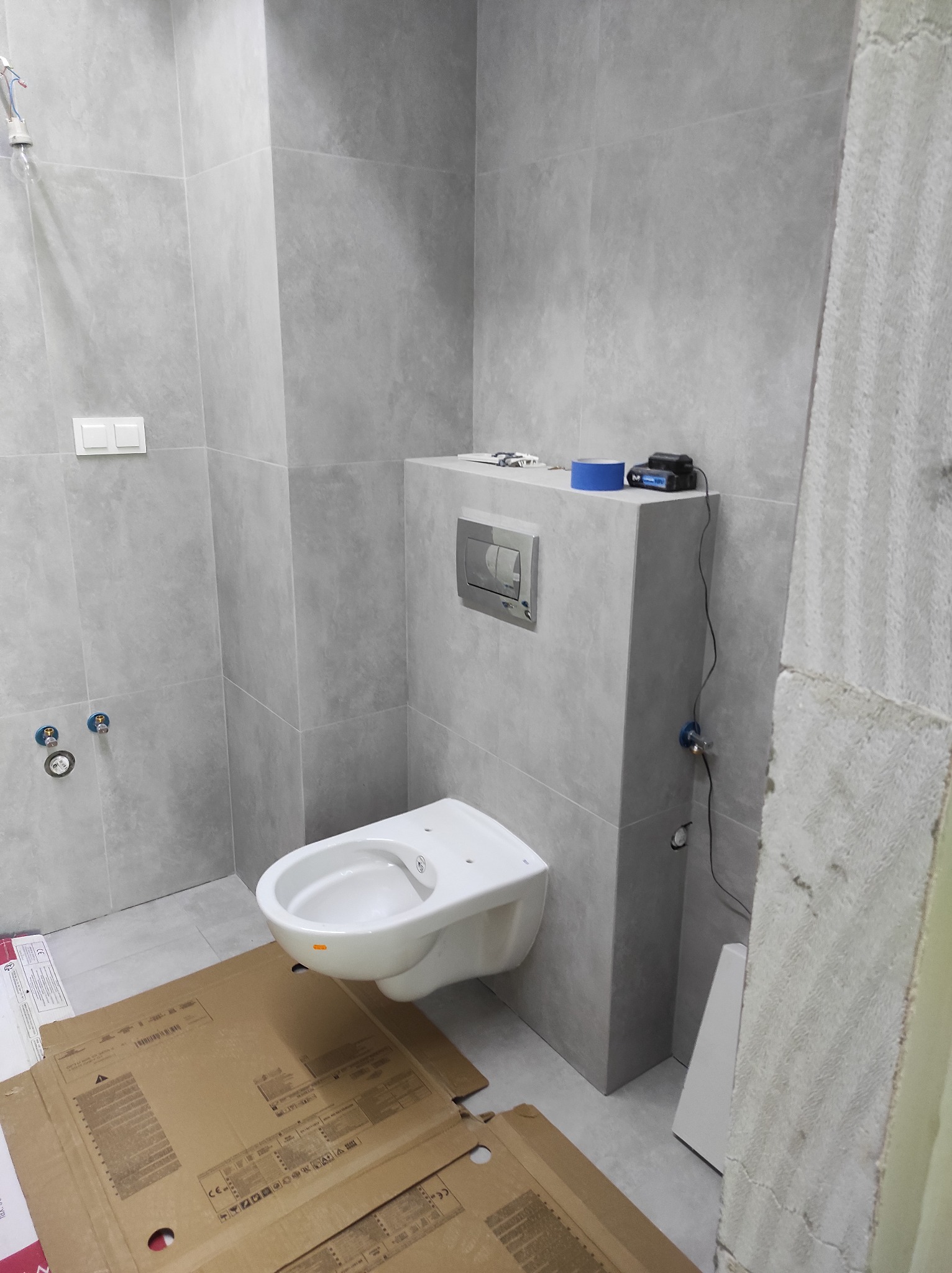 Szara łazienka w trakcie remontu: podwieszana toaleta, płytki imitujące beton, kartony na podłodze. Widoczne narzędzia i akcesoria instalacyjne.