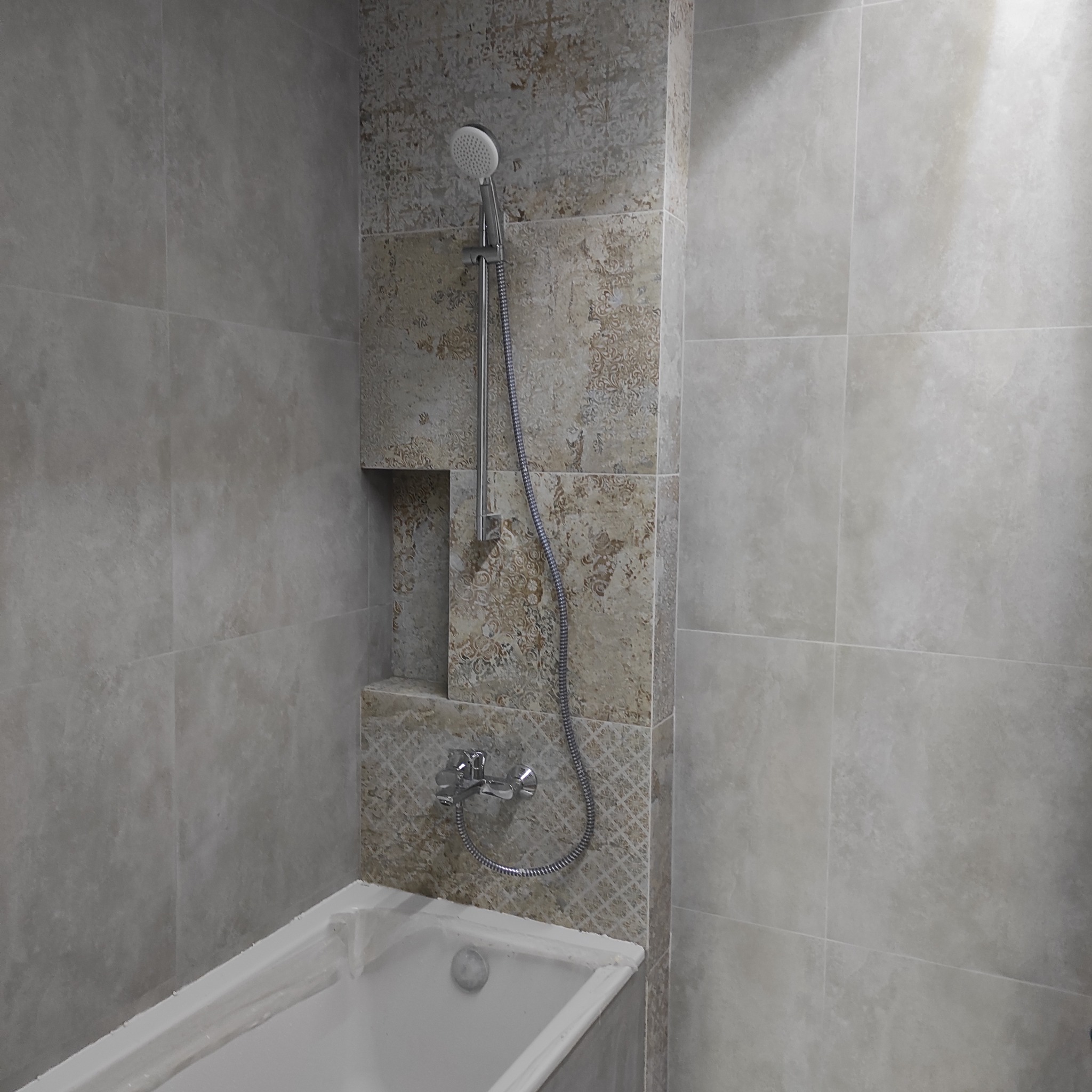 Wykończona łazienka z wanną i prysznicem. Ściany wyłożone płytkami w odcieniach szarości i beżu, z dekoracyjnym wzorem w strefie prysznica i wnęką na kosmetyki.