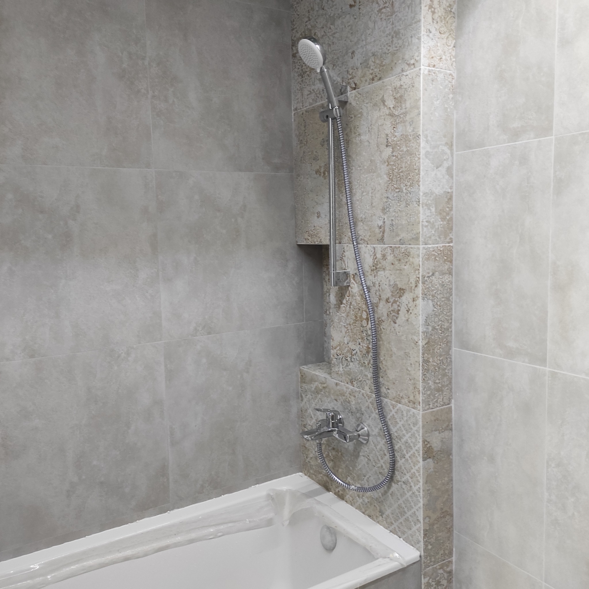 Wykończona łazienka z beżowo-szarą glazurą, chromowaną armaturą i wnęką prysznicową. Widoczna wanna zabezpieczona folią ochronną.