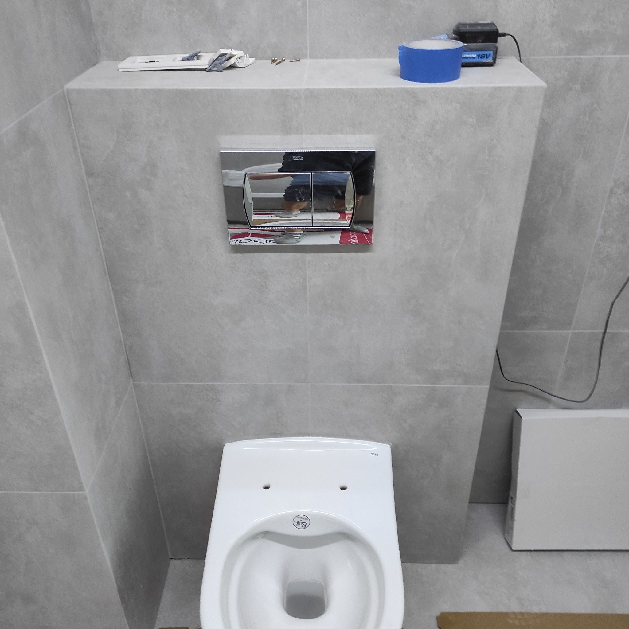 Szara łazienka z podtynkową toaletą Roca i chromowanym przyciskiem spłukującym. Na górze widoczne narzędzia i niebieska taśma malarska. Minimalistyczny design.