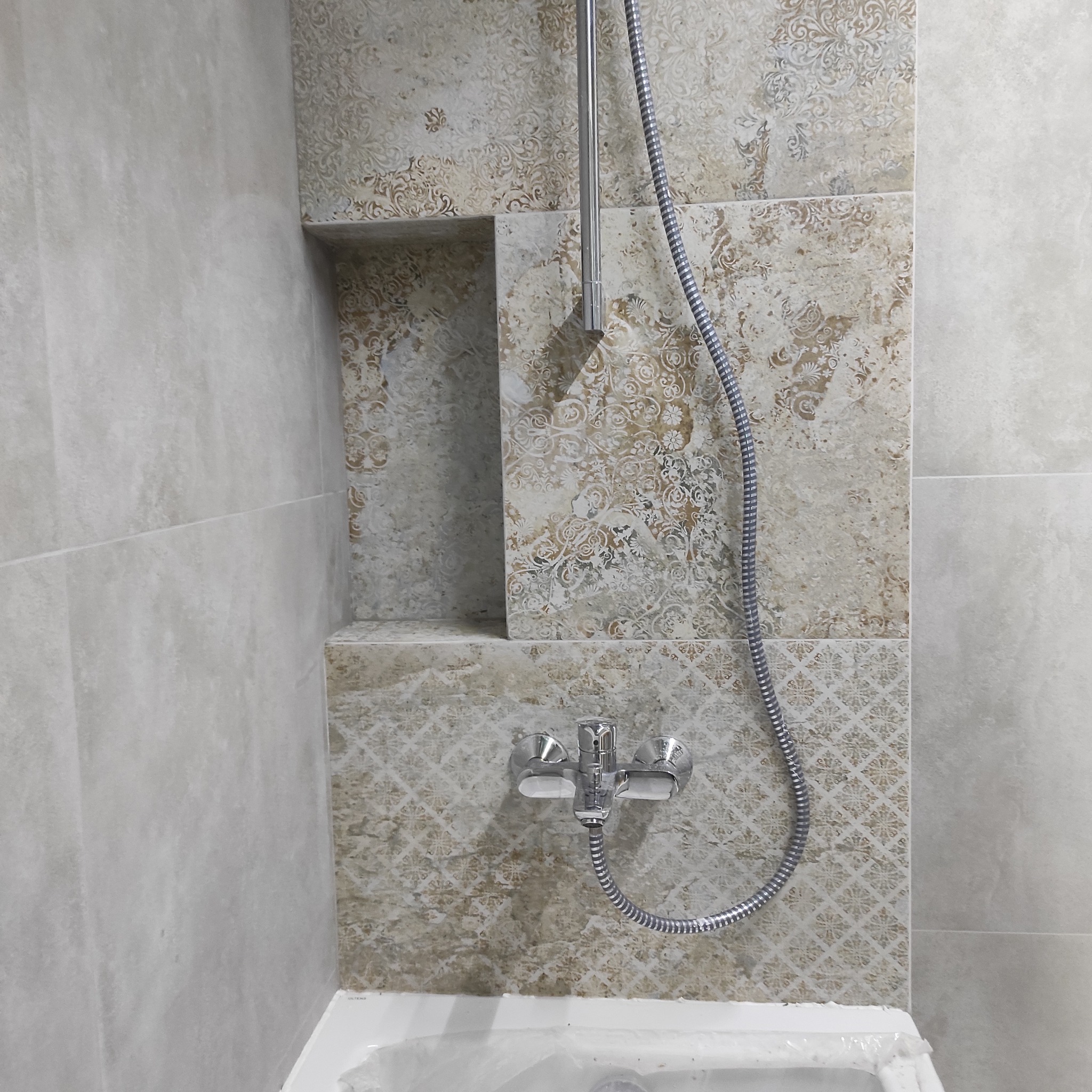 Wykończona ściana w łazience z beżowymi płytkami o wzorzystej fakturze, wnęka i bateria prysznicowa. Częściowo widoczna wanna zabezpieczona folią.