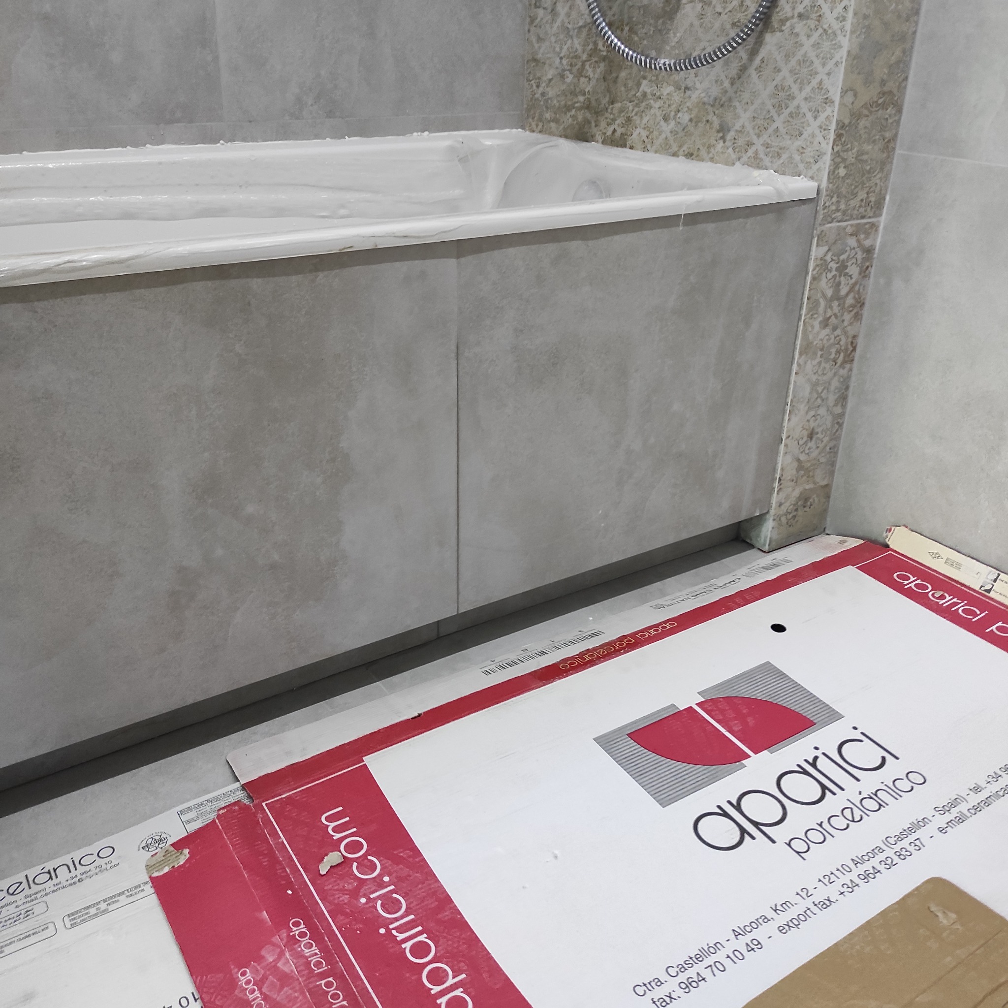 Wykończona obudowa wanny z szarych płytek, widoczny fragment kabiny prysznicowej i karton z logo producenta płytek. Detal zrealizowanej łazienki.