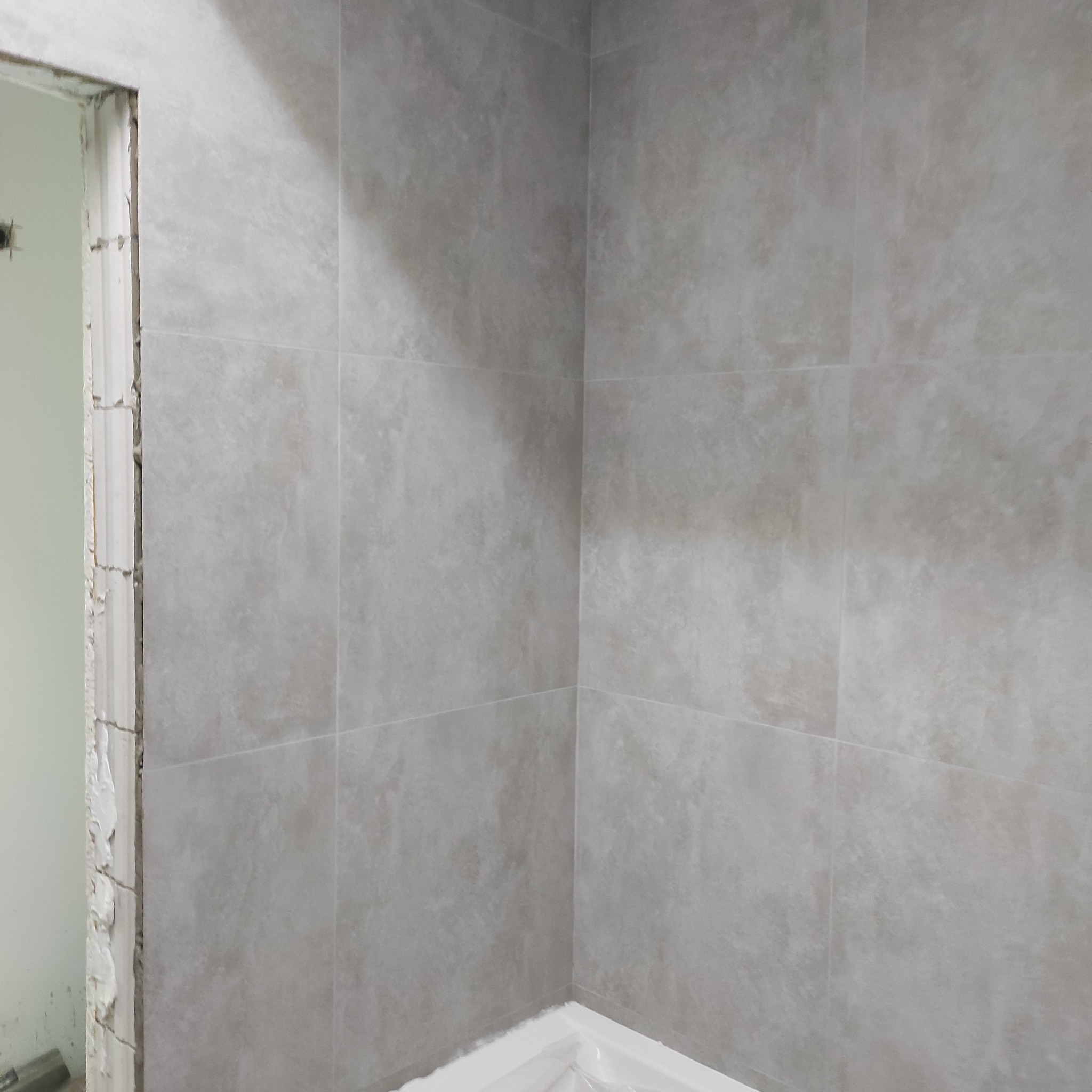 Wykończona ściana łazienki z szarych, matowych płytek wielkoformatowych. Widoczny fragment obudowy wanny i surowy otwór drzwiowy z resztkami zaprawy.