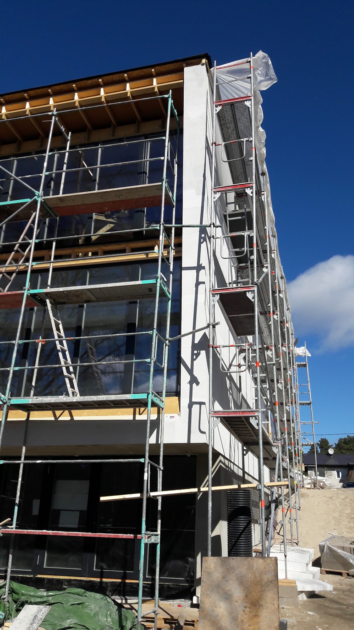 Elewacja domu w trakcie prac wykończeniowych, widoczne rusztowania i fragment ocieplenia. Błękitne niebo w tle.