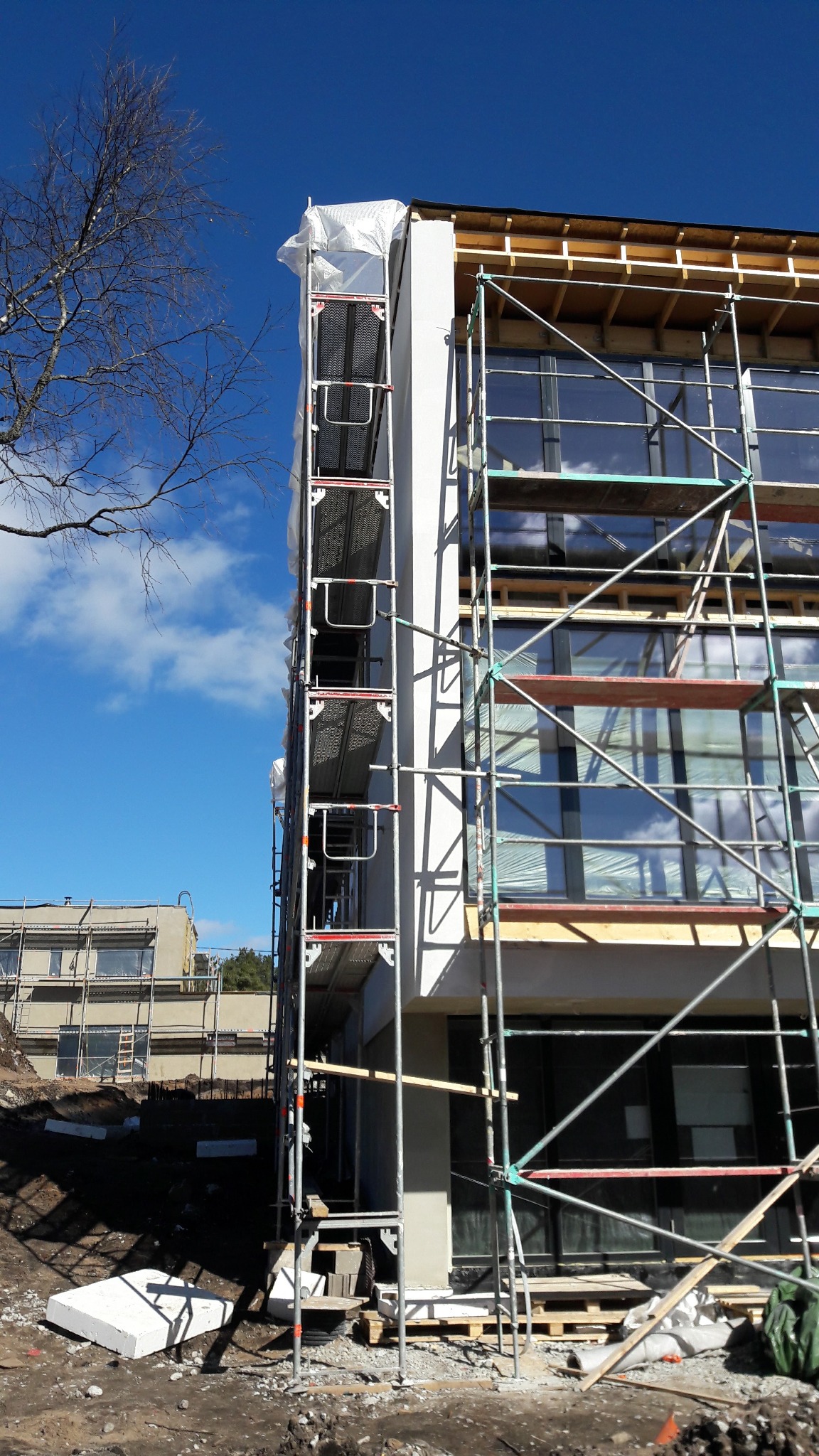 Budynek w trakcie ocieplania, osłonięty folią i otoczony rusztowaniami. Widoczne okna i fragment drugiego budynku w tle, błękitne niebo.