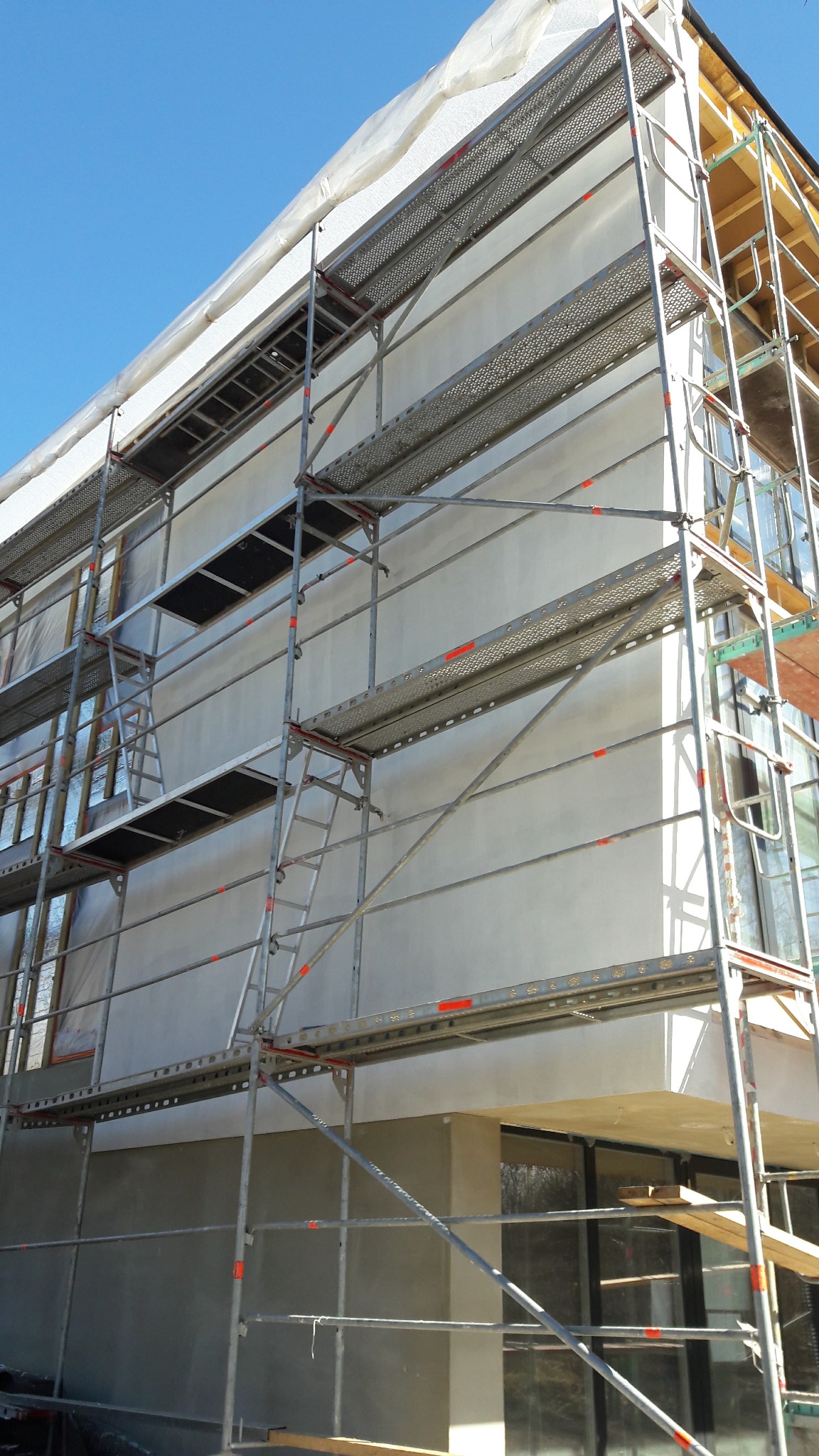 Elewacja budynku w trakcie tynkowania, widoczne rusztowania i zabezpieczone okna folią. Świeży tynk na ścianie, błękitne niebo w tle. Ujęcie z dołu.