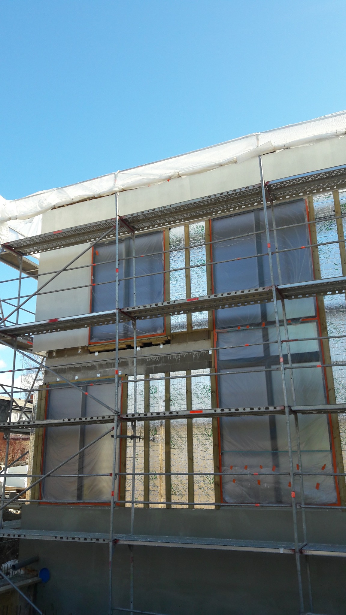 Ocieplanie ściany budynku z zewnątrz, widoczne rusztowanie, folia ochronna i warstwy izolacyjne. Prace w toku, jasne niebo w tle.