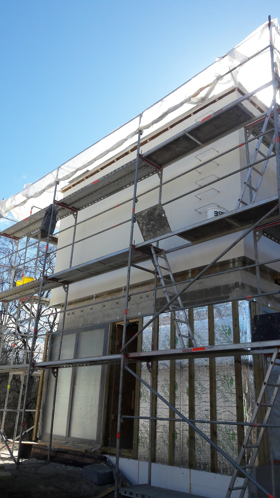 Ocieplanie budynku z zewnątrz, widoczne rusztowania, ocieplona ściana budynku, folia izolacyjna i fragment błękitnego nieba. Prace w toku, etap ocieplania.