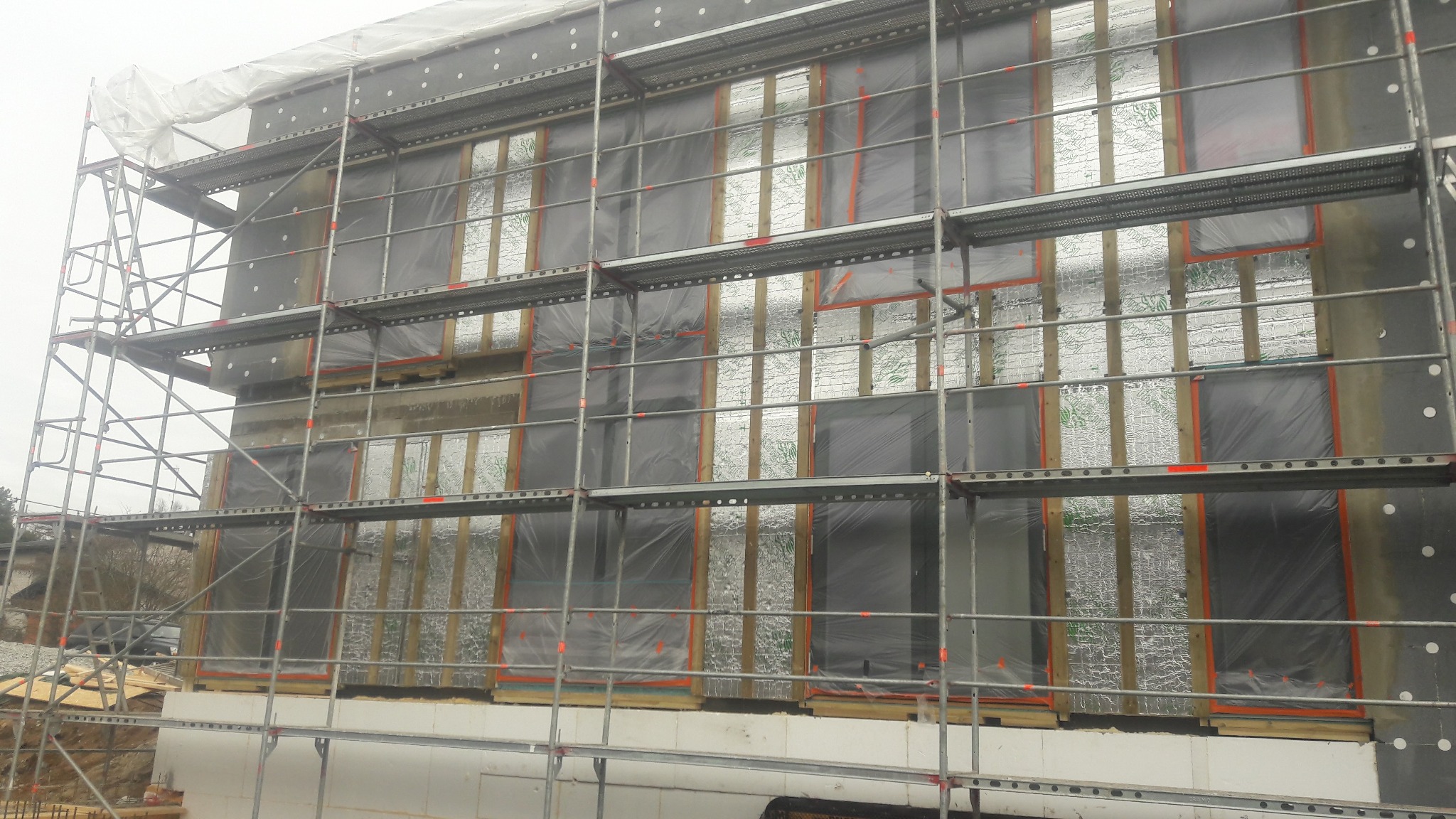 Budynek w trakcie ocieplania, widoczne rusztowania, izolacja termiczna i folia ochronna. Prace na wysokości, etap ocieplania ścian zewnętrznych.