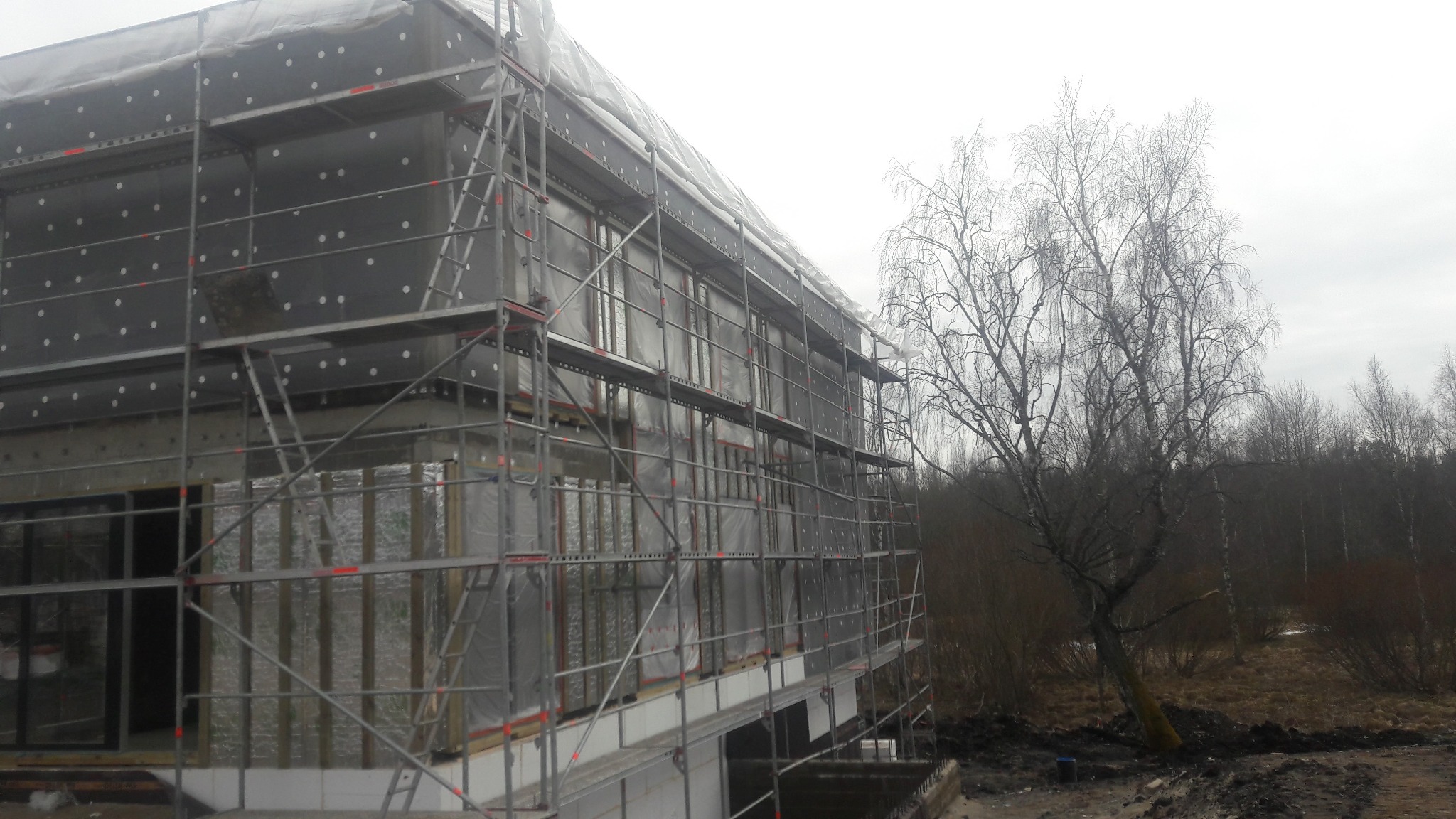 Dom w trakcie ocieplania, widoczne rusztowania i materiały izolacyjne. Na pierwszym planie drzewo bez liści, pochmurne niebo w tle. Prace w toku.