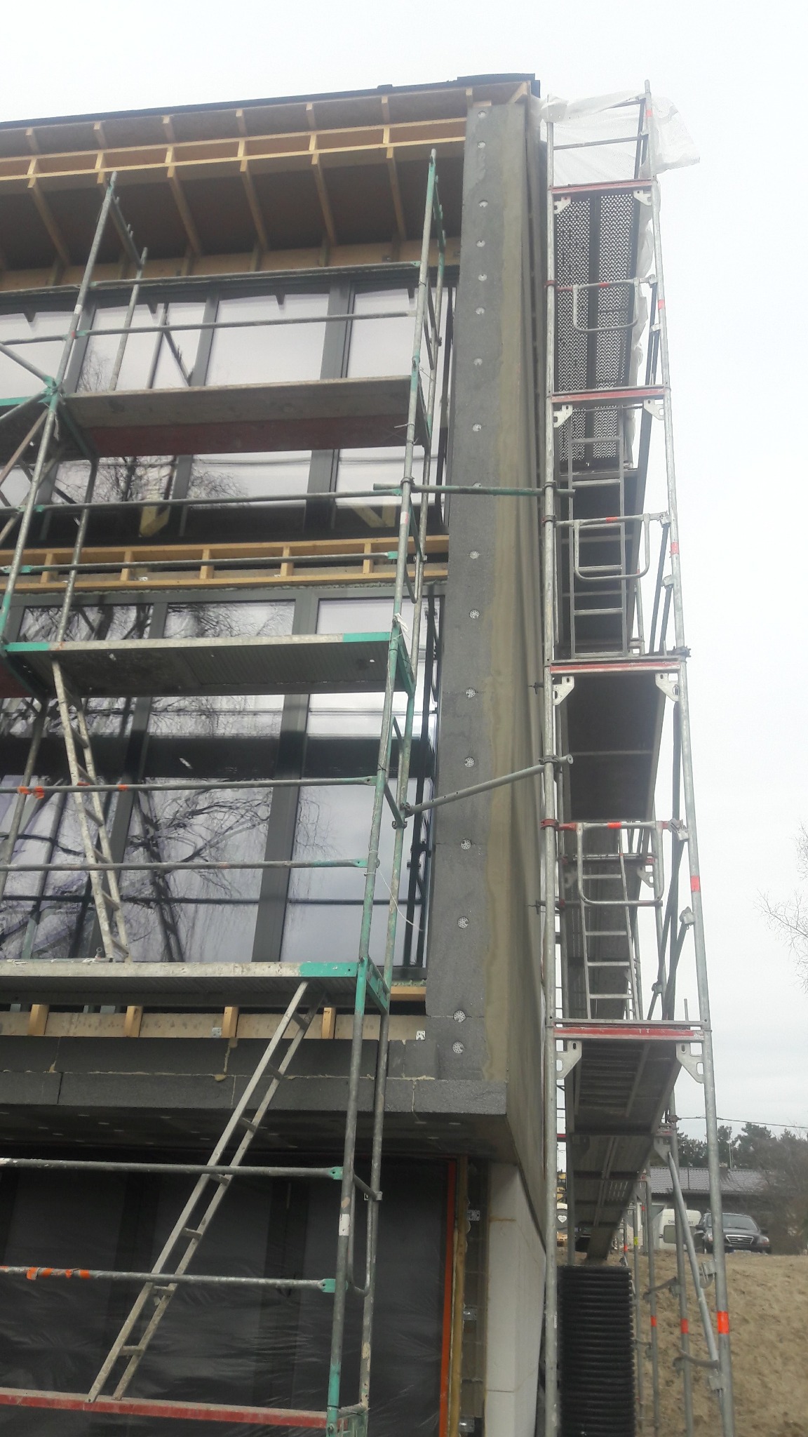 Ocieplanie budynku z użyciem rusztowań. Widoczne płyty izolacyjne i okna. Prace w toku, elewacja w trakcie wykonywania. Szare niebo w tle.