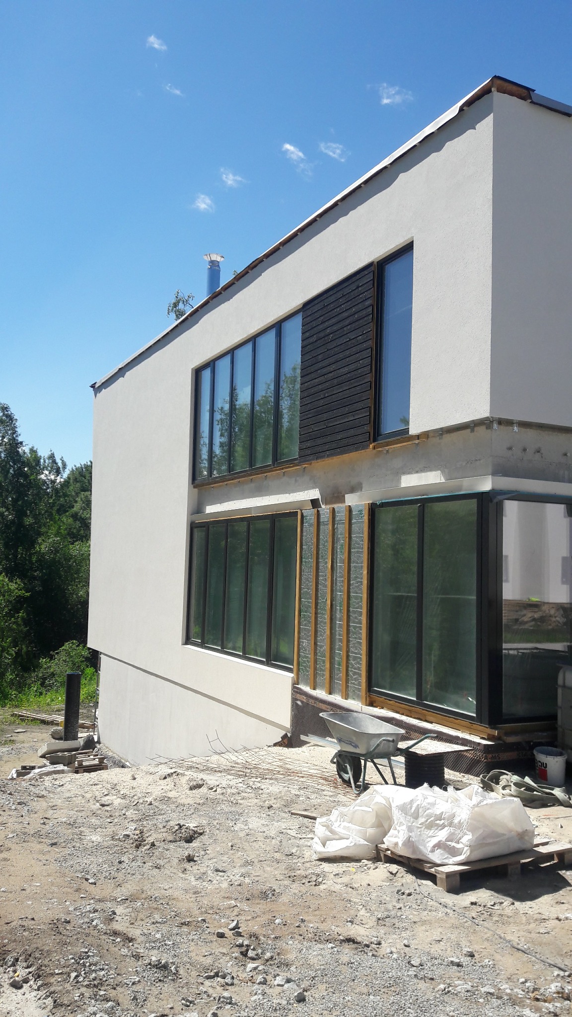 Nowoczesny dom z oknami w trakcie wykończenia elewacji. Widoczne rusztowania, worki z materiałami budowlanymi i taczka. Jasne tynki i drewniane elementy.