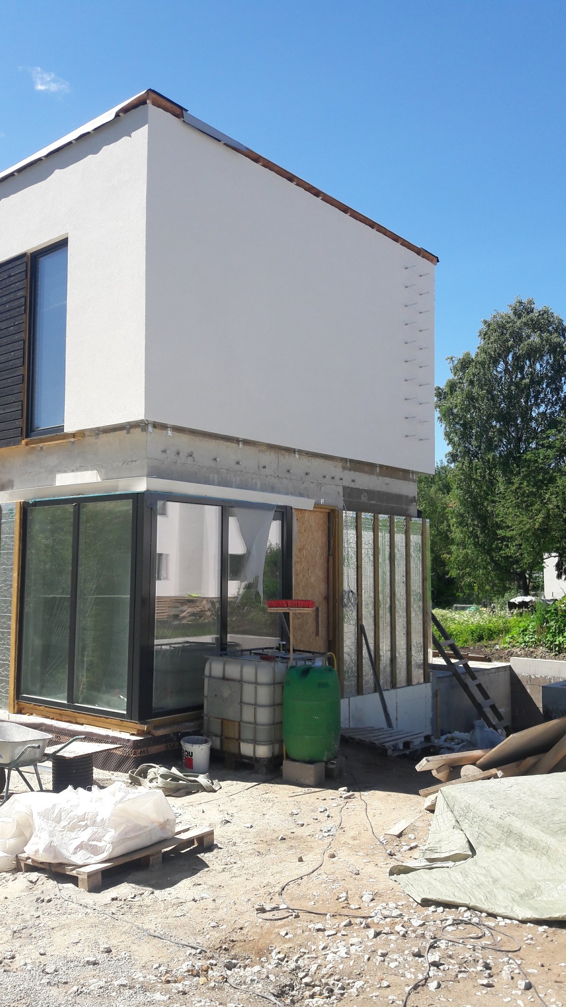 Ocieplony budynek w trakcie prac wykończeniowych. Widoczne materiały izolacyjne, okna z ciemnymi ramami i biała elewacja. Na pierwszym planie zbiorniki na wodę i gruz.