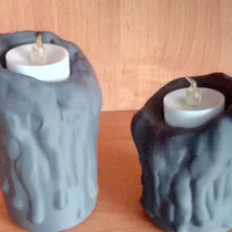 Dwa ozdobne świeczniki w kształcie nieregularnych brył z szarego tworzywa, zawierające białe, elektryczne świece imitujące płomień, ustawione na drewnianej półce.