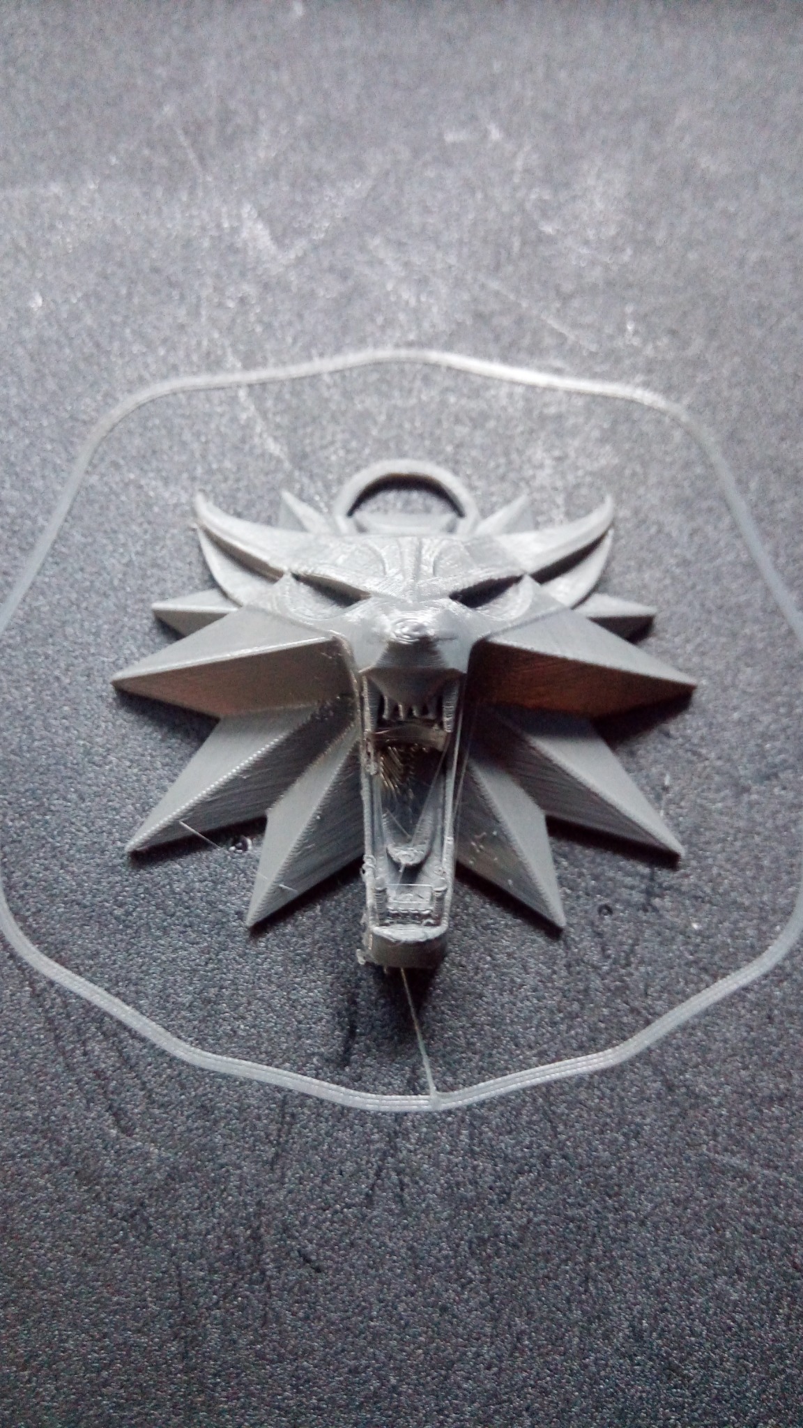 Szary medalion wilka z otwartą paszczą, wydrukowany w 3D na szarej teksturowanej powierzchni, widoczne niedoskonałości druku.