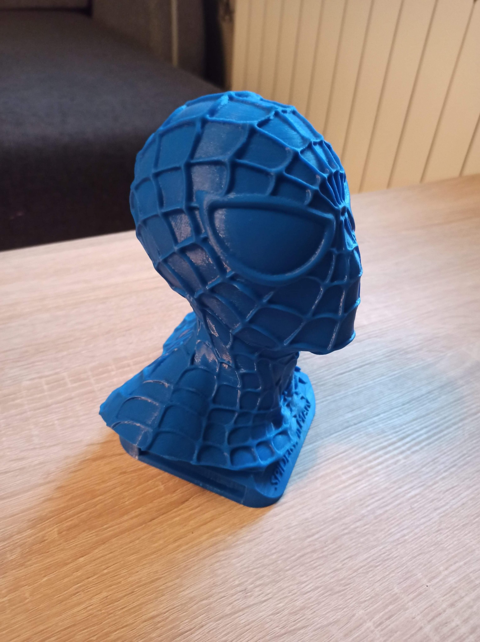 Niebieska, trójwymiarowa głowa Spidermana wydrukowana na drukarce 3D, umieszczona na drewnianym stole.