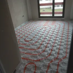 Pomieszczenie w trakcie montażu ogrzewania podłogowego: rury grzewcze w kolorze pomarańczowym ułożone na białej izolacji systemowej z wypustkami, widoczne okno i surowe ściany.