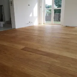 Wykończona podłoga z desek drewnianych o ciepłym, miodowym odcieniu w jasnym, minimalistycznym wnętrzu z białymi ścianami i dużym oknem balkonowym.